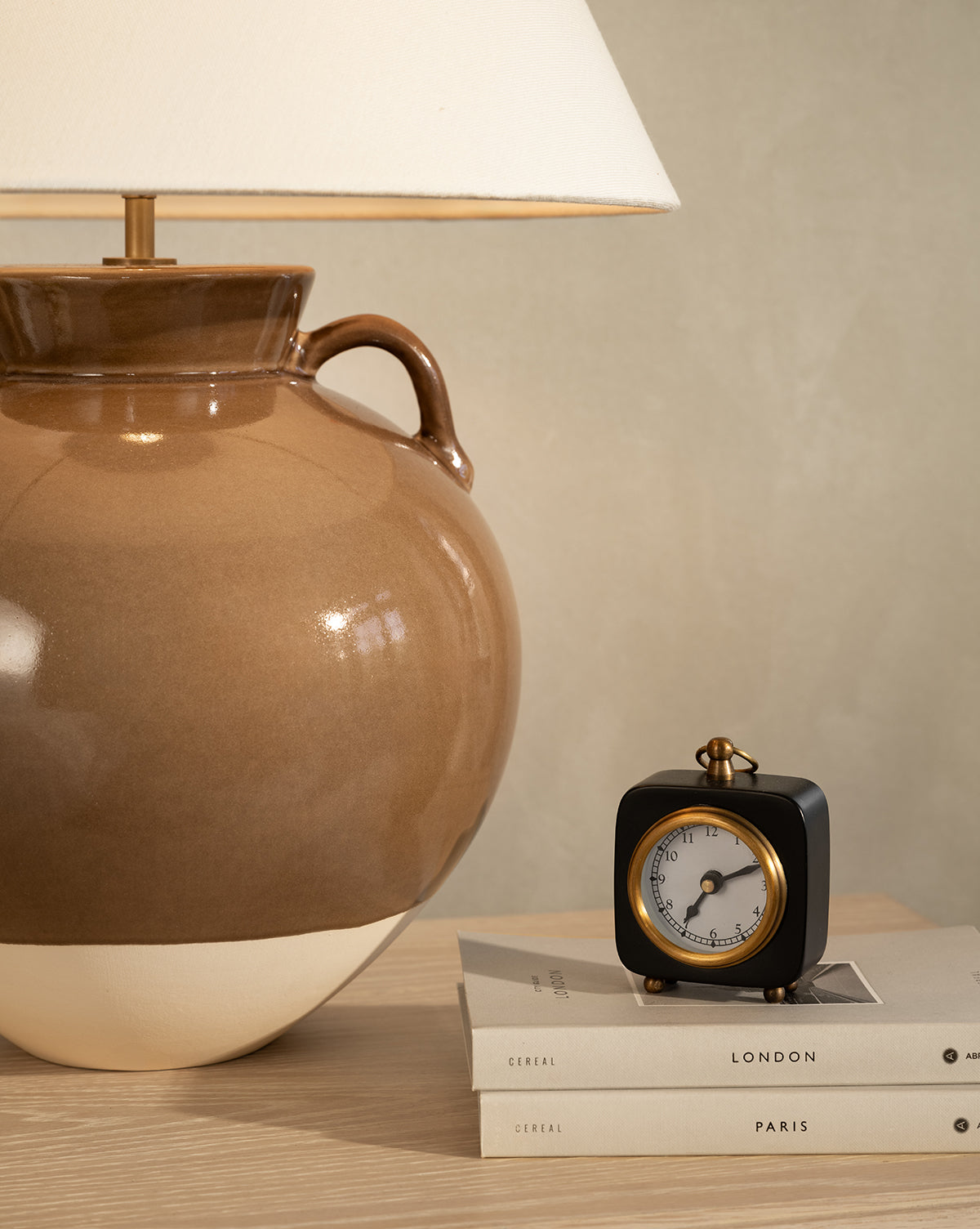 A brown ceramic lamp and the Kya Clock sit on books labeled Cereal, London, and Paris atop a light wooden nightstand against a beige wall, by Vendixs