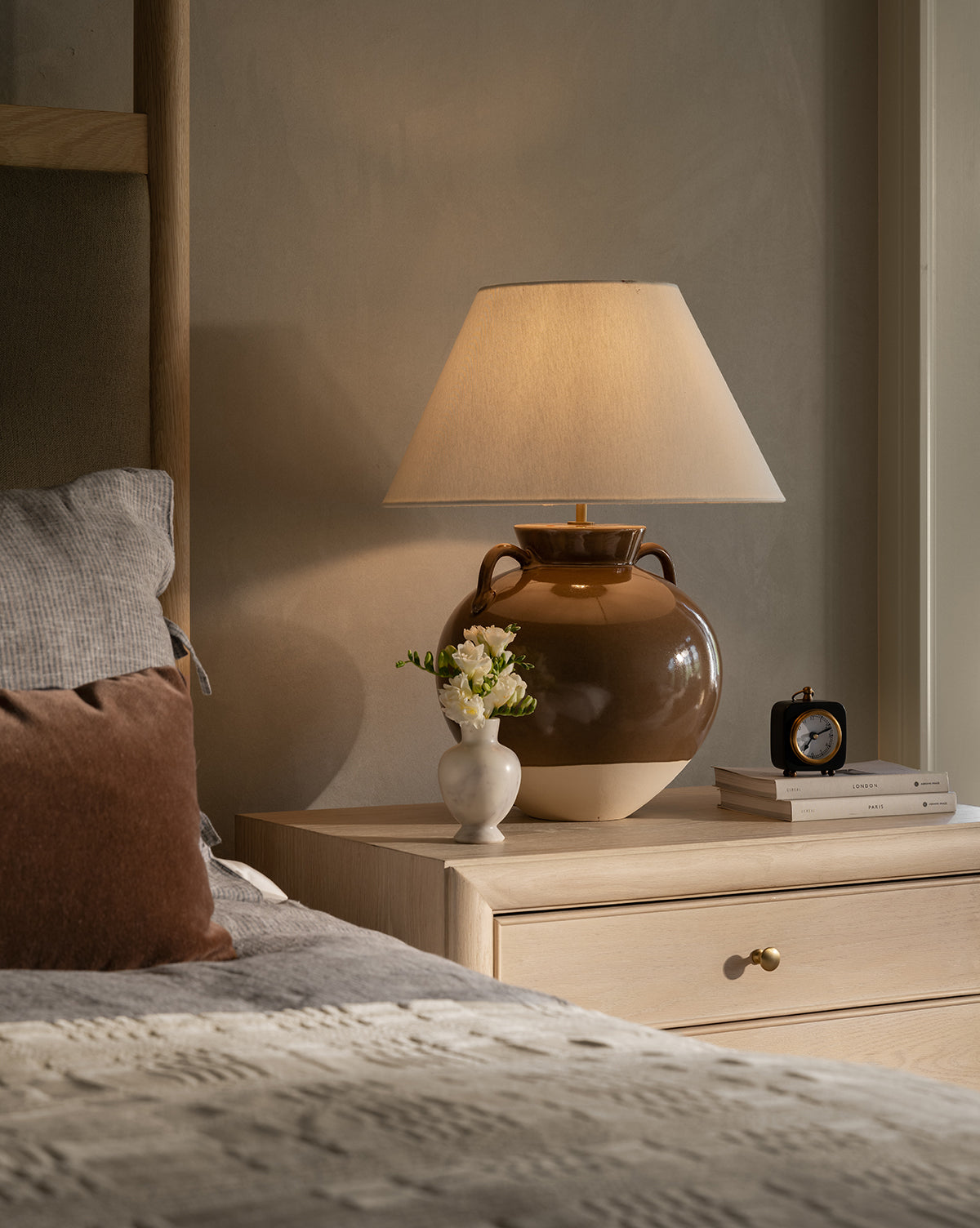 A cozy bedside scene features the Amphora Table Lamp, a vase of white flowers, books, and a black clock on a light wood nightstand beside a bed with neutral bedding by Vendixs