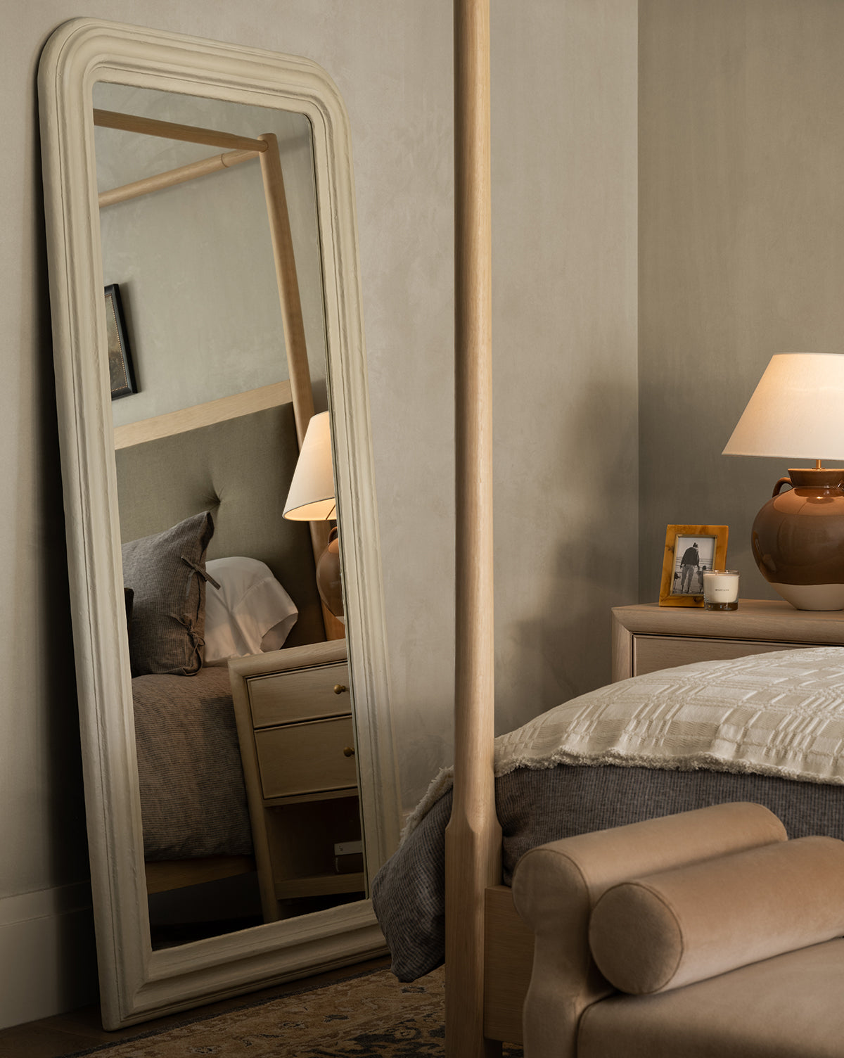 A cozy bedroom features a Breslin Floor Mirror reflecting the bed, nightstand, and lamp. A beige knit blanket, tan leather bench, and brown lamp with a photo frame sit on the nightstand near plaster-finished walls by Vendixs