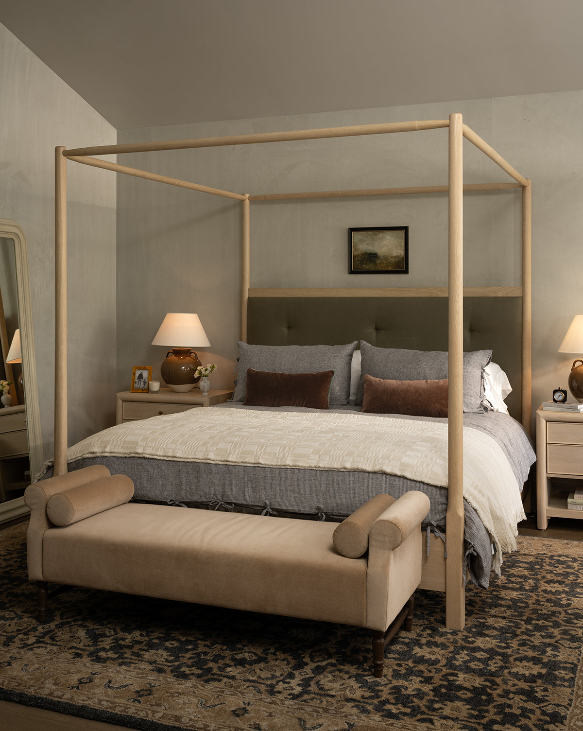 A cozy bedroom featuring a wooden four-poster bed with Makerspalms Chamberlain Coverlet in gray and cream, brown accent pillows, two bedside tables with lamps, a cushioned bench at the foot, and a large patterned rug on the floor. Vendixs