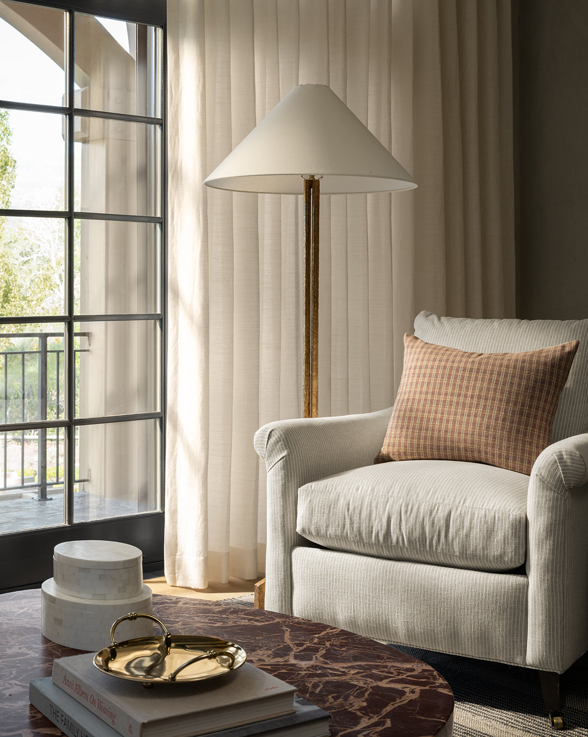 A cozy living room corner features the Reedley Lounge Chair by Rowe Fine Furniture, Inc., a brown checkered pillow, gold floor lamp, sheer curtains, and a marble coffee table with books and decorative boxes near a large window—finished with an accent from Vendixs