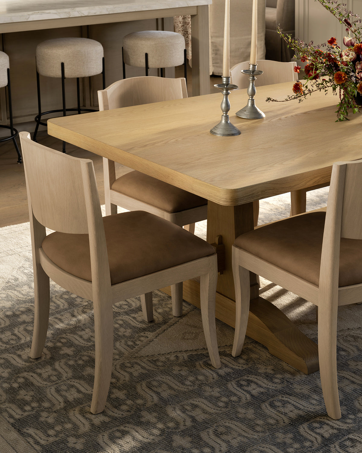 The Wilton Dining Table with four light wood chairs with brown cushions, two silver candlesticks, and a floral arrangement sits on a patterned rug in a modern dining area by McGee & Co.