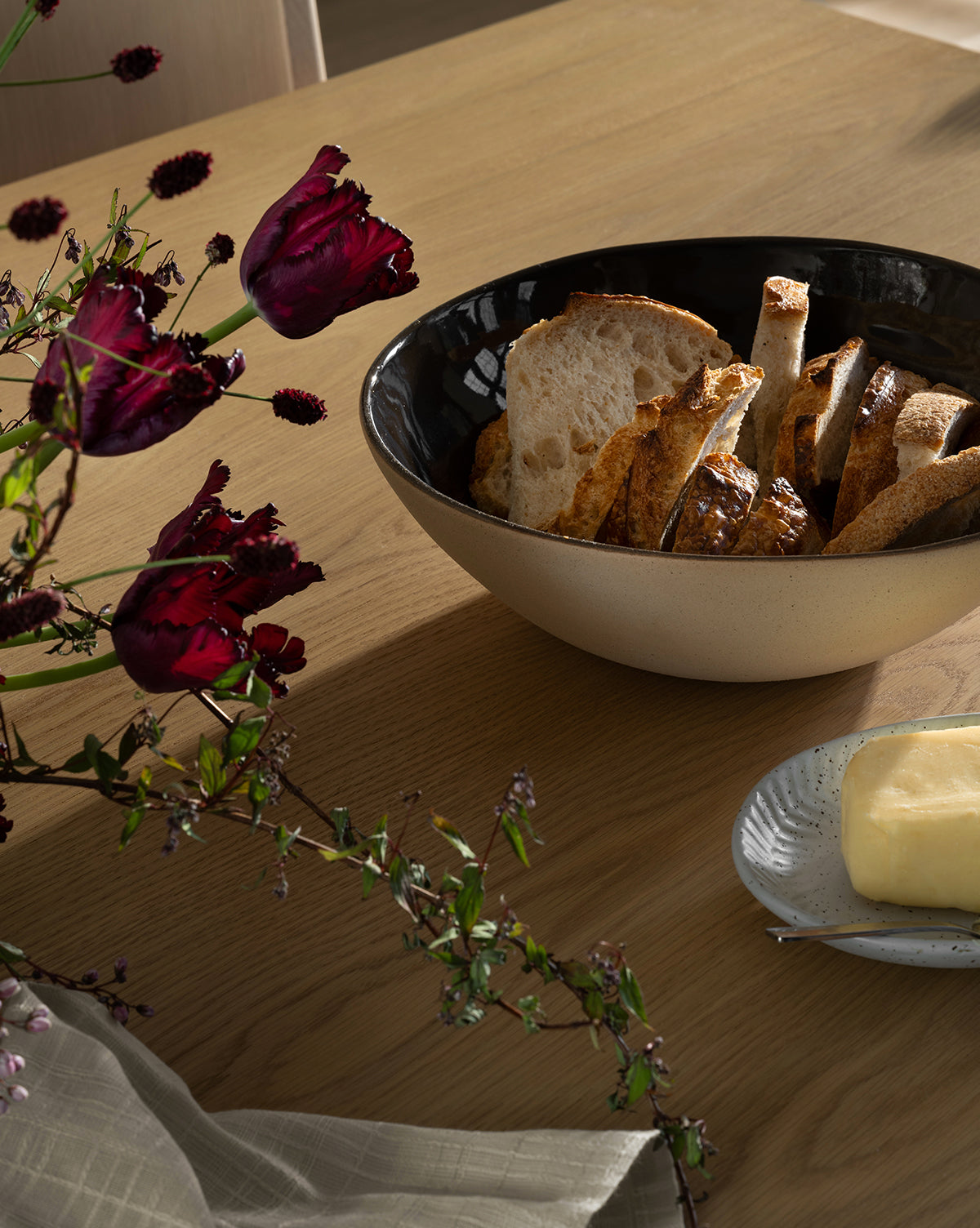The Joanne Serving Bowl, filled with sliced artisan bread, sits on a wooden table next to butter, dark red flowers, and a folded napkin by Vendixs