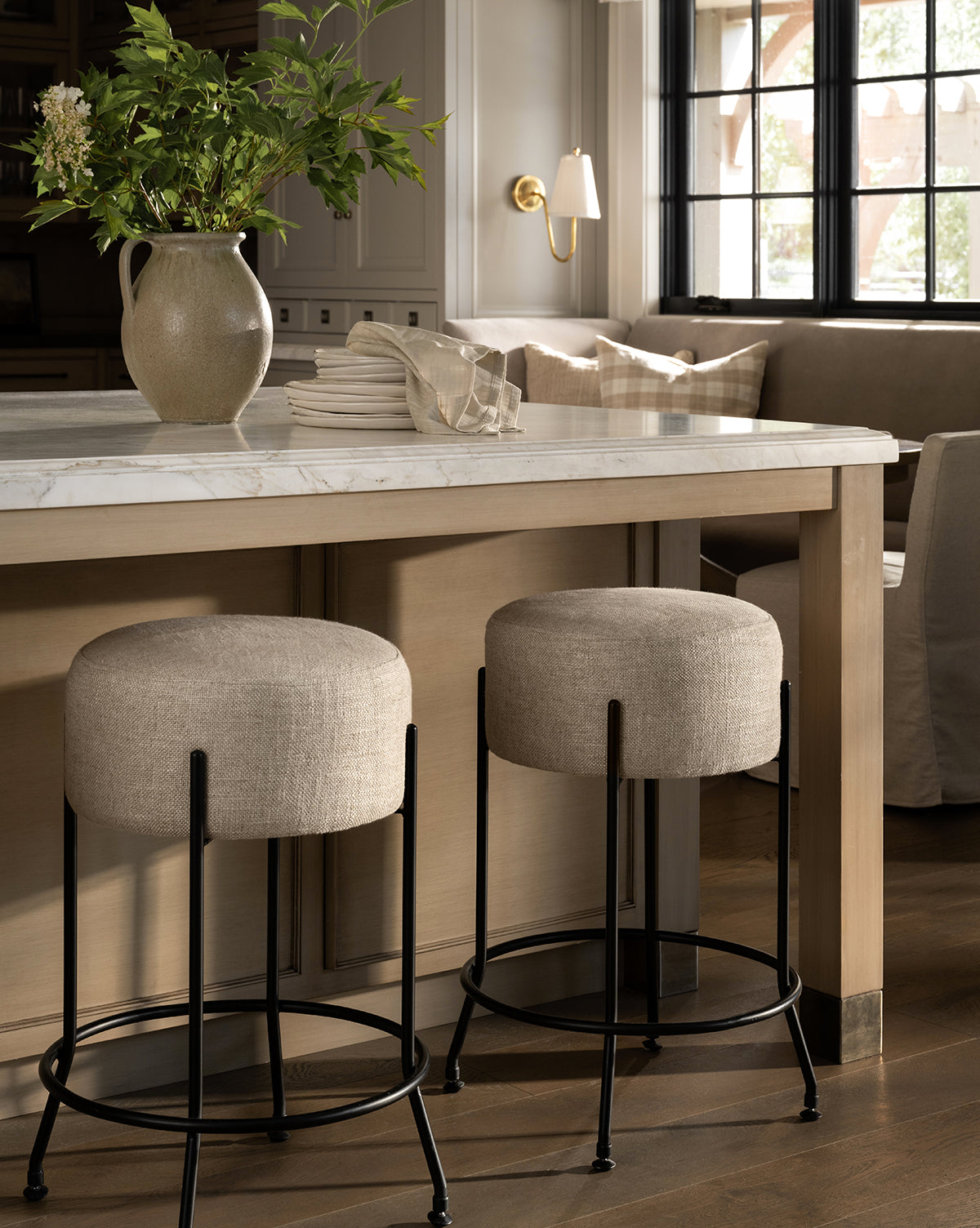 Certainly! Here’s your revised sentence with Vendixs blended at the end: A modern kitchen showcases a marble countertop island with a beige vase of greenery, stacked white plates, and two Makerspalm Evander Counter Stools—industrial-inspired with black metal legs—plus a window seat in the background, all styled by Vendixs