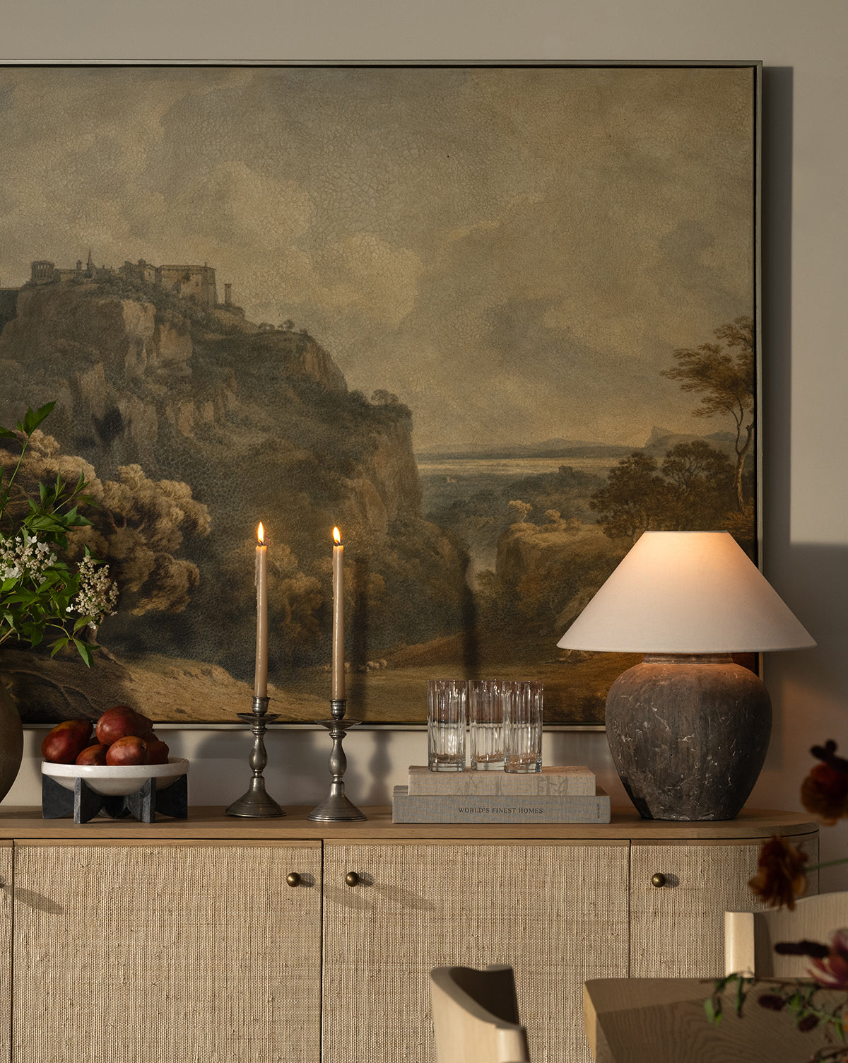 A cozy sideboard holds a rustic table lamp, two lit candles, stacked books, apples in a bowl, glasses, and a vase of flowers beneath the dramatic Overlooking Tivoli landscape painting of a cliffside village by Vendixs