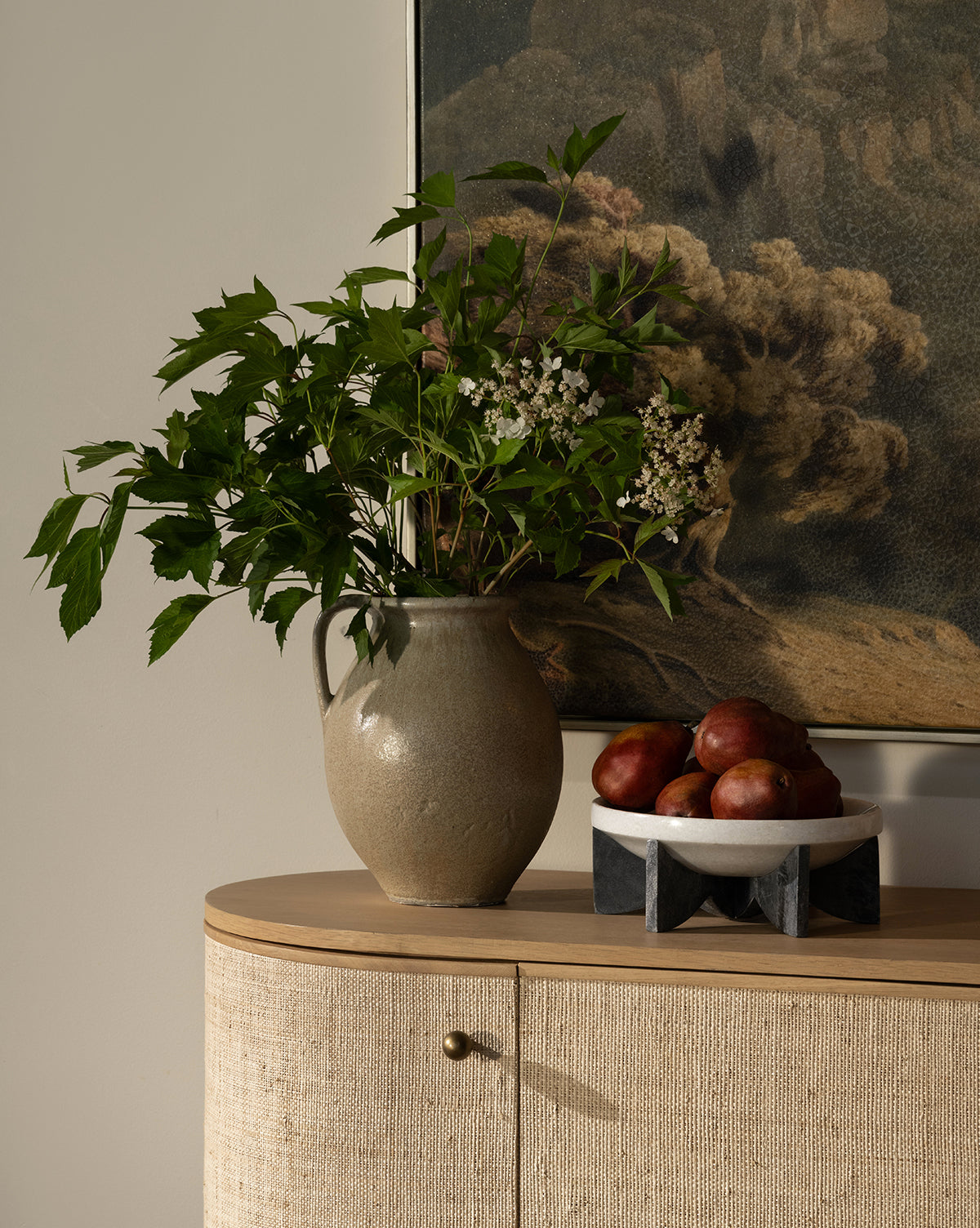 A stoneware jug with green leafy branches and white flowers sits on a light wood cabinet next to the Mahoney Bowl filled with red apples, by Vendixs A landscape painting hangs on the wall in the background.