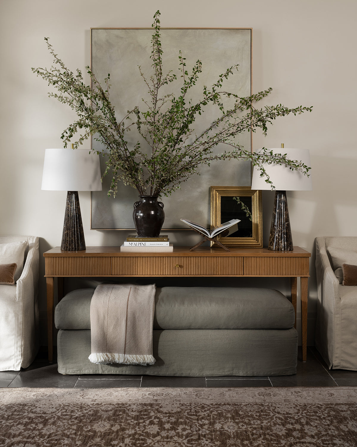 A console table with a large black vase of leafy branches, two table lamps, books, and a framed picture sits between sofas. The Macy Slipcover Ottoman, with a folded throw on top, is placed beneath the table by McGee & Co.