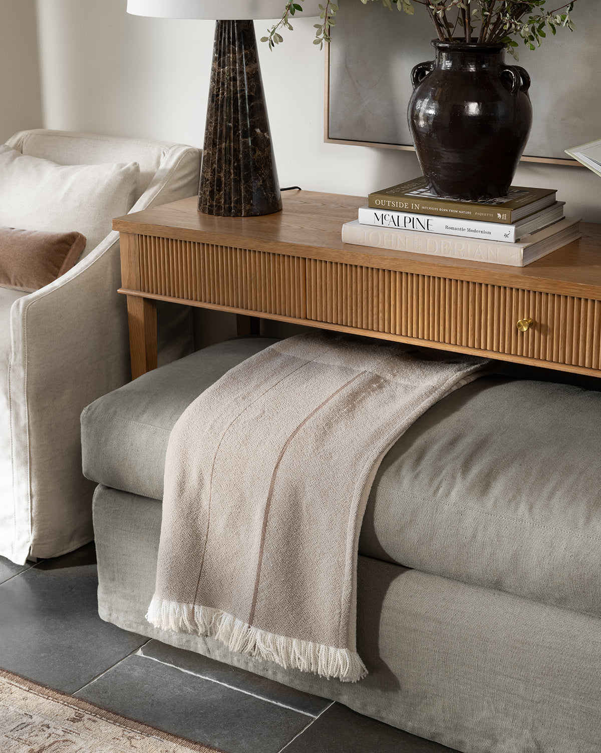 A cozy neutral-toned living room corner features a wooden console table with a dark vase, books, and a marble lamp. Beneath it, a cushioned bench displays the Alex Plaid Throw by ILA HOME FASHIONS in light beige for added texture. Styled by Vendixs