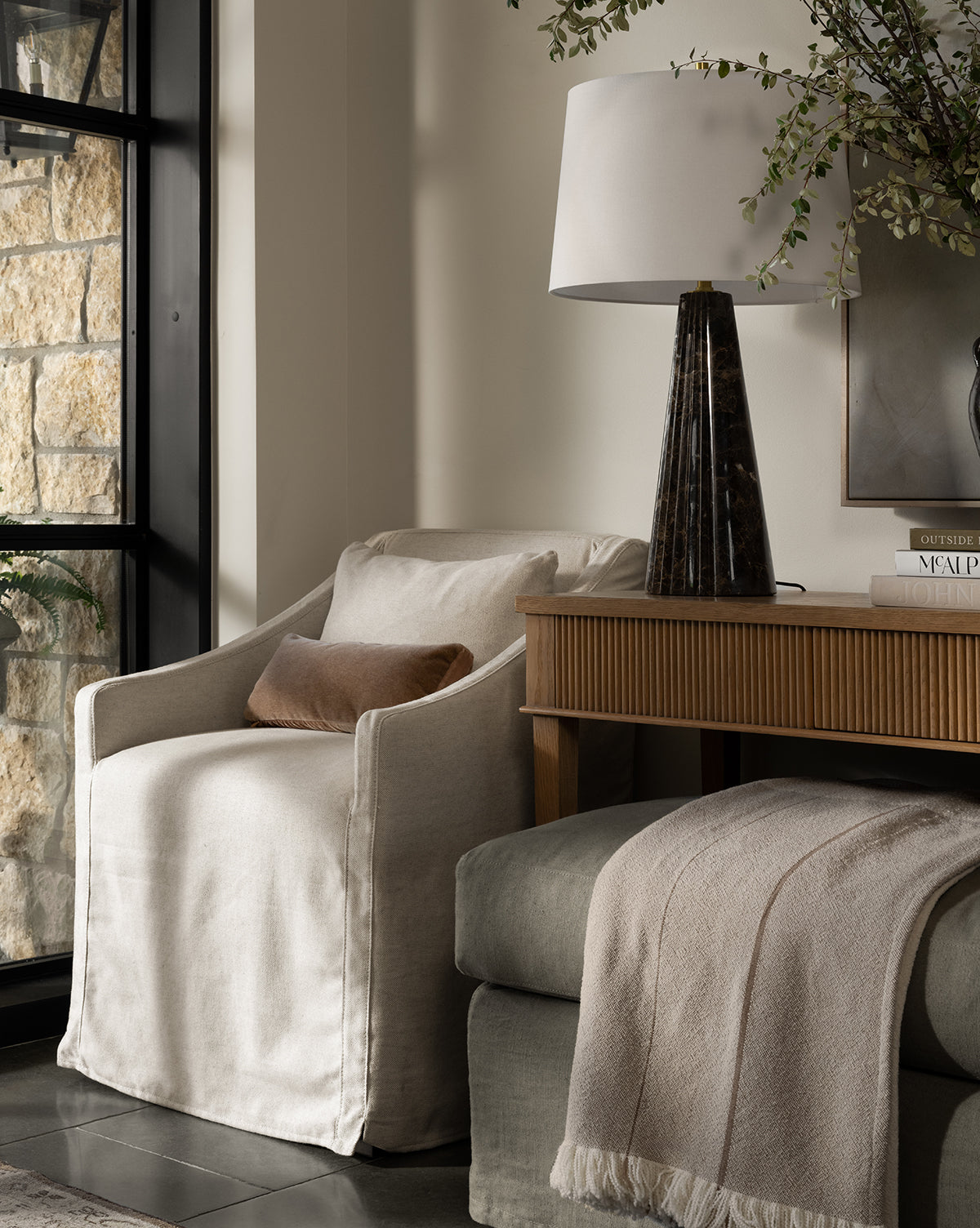 A cozy corner features the Olivier Slipcover Dining Arm Chair in cream with a brown feather and down cushion, paired with a wooden console table, dark lamp, books, a light throw on an ottoman, greenery in a vase, and a large stone-and-black-framed window, by McGee & Co.
