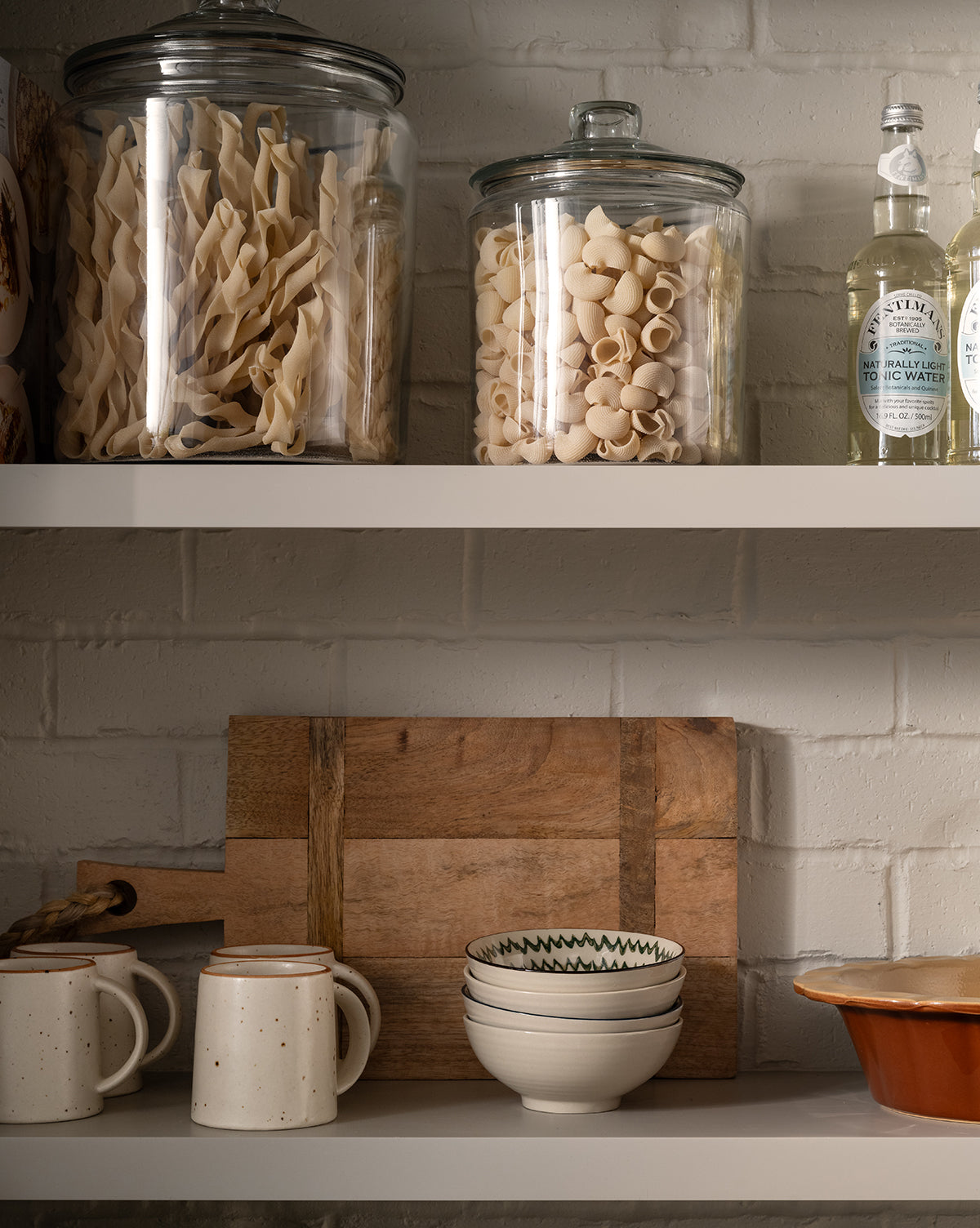 Two glass jars of pasta and bottles sit on an upper shelf; below, ceramic mugs, a wooden cutting board, stacked bowls including the Mattia Hand-Painted Bowl with floral patterns, and a ceramic dish rest against a white brick wall by Vendixs