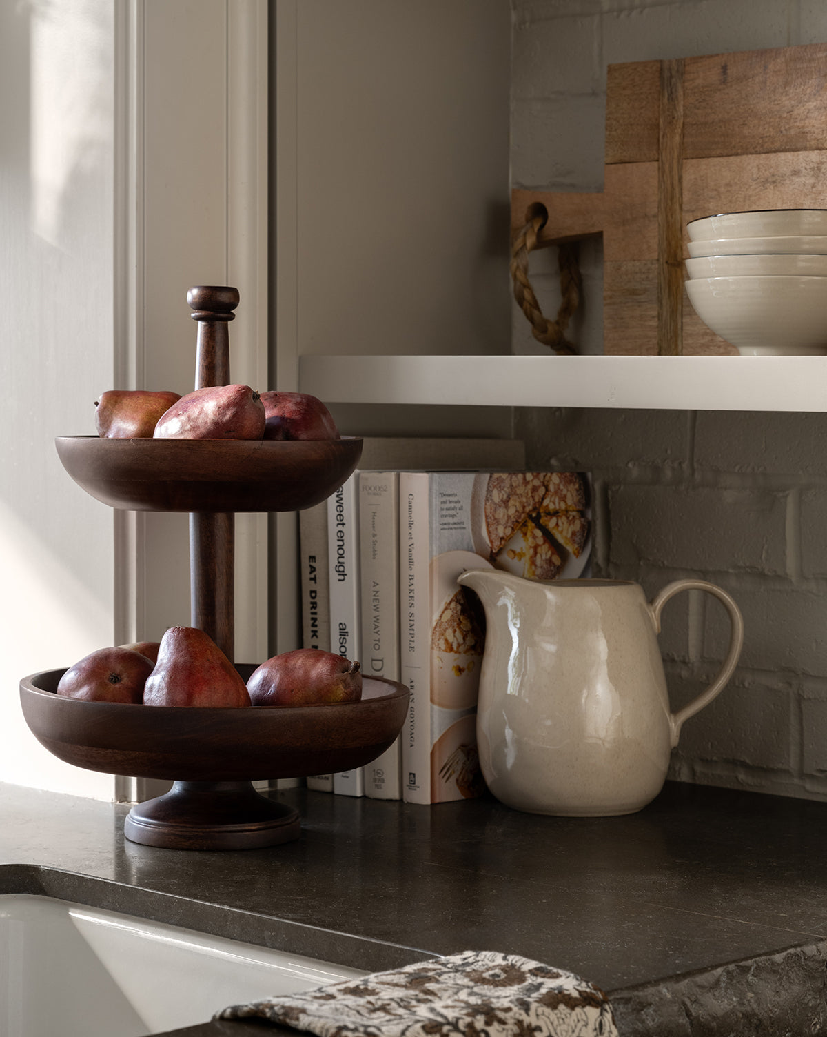 A Winward Tiered Pedestal in dark wood holds red pears on a kitchen counter beside stacked cookbooks, a white ceramic pitcher, and neatly arranged bowls displayed on the shelf above, by Vendixs