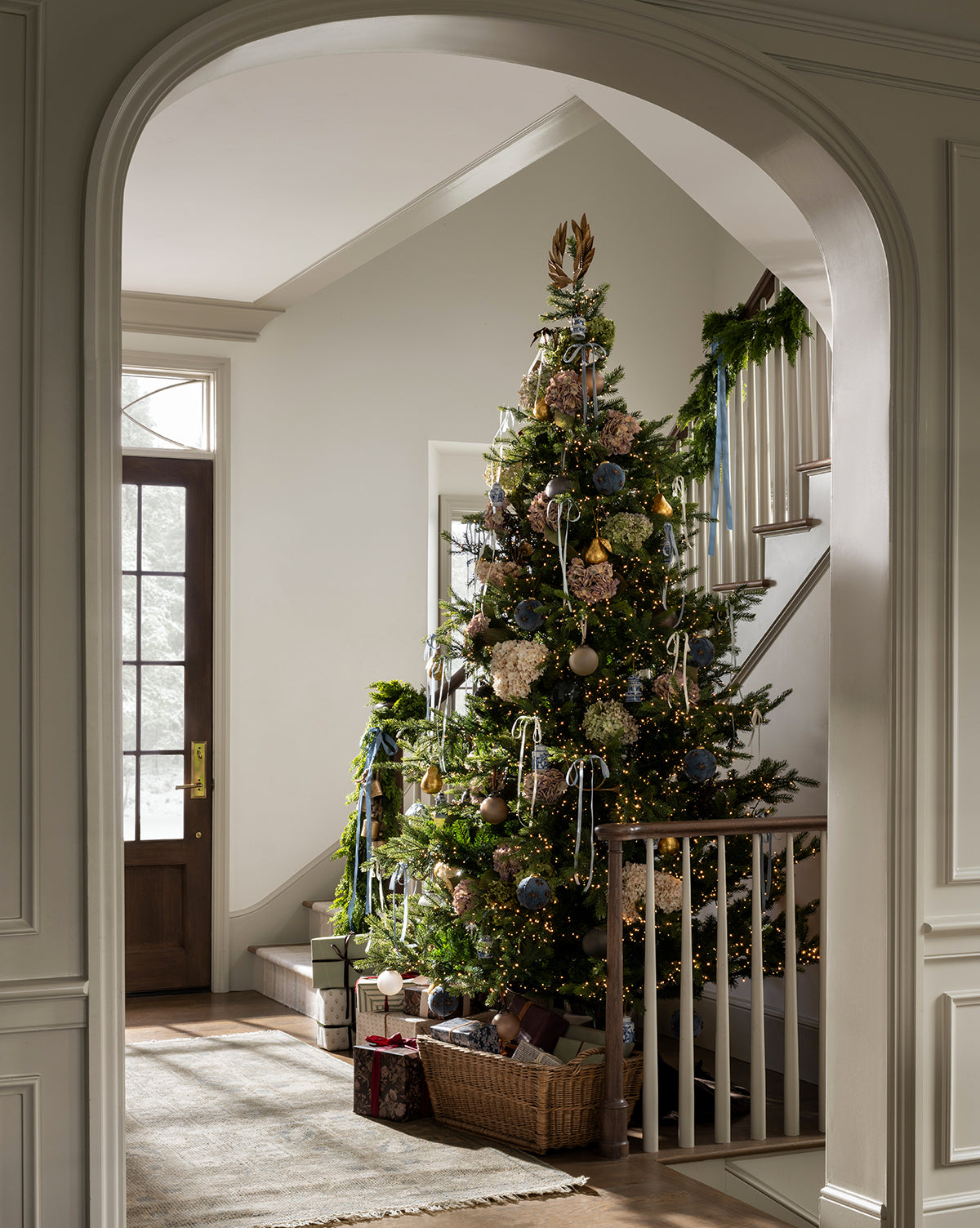 A 9' Norway Spruce Pre-Lit Faux Tree decorated with ornaments, ribbons, and lights stands by a staircase in a bright entryway. Wrapped gifts sit below as greenery lines the banister and natural light fills the space, by Vendixs