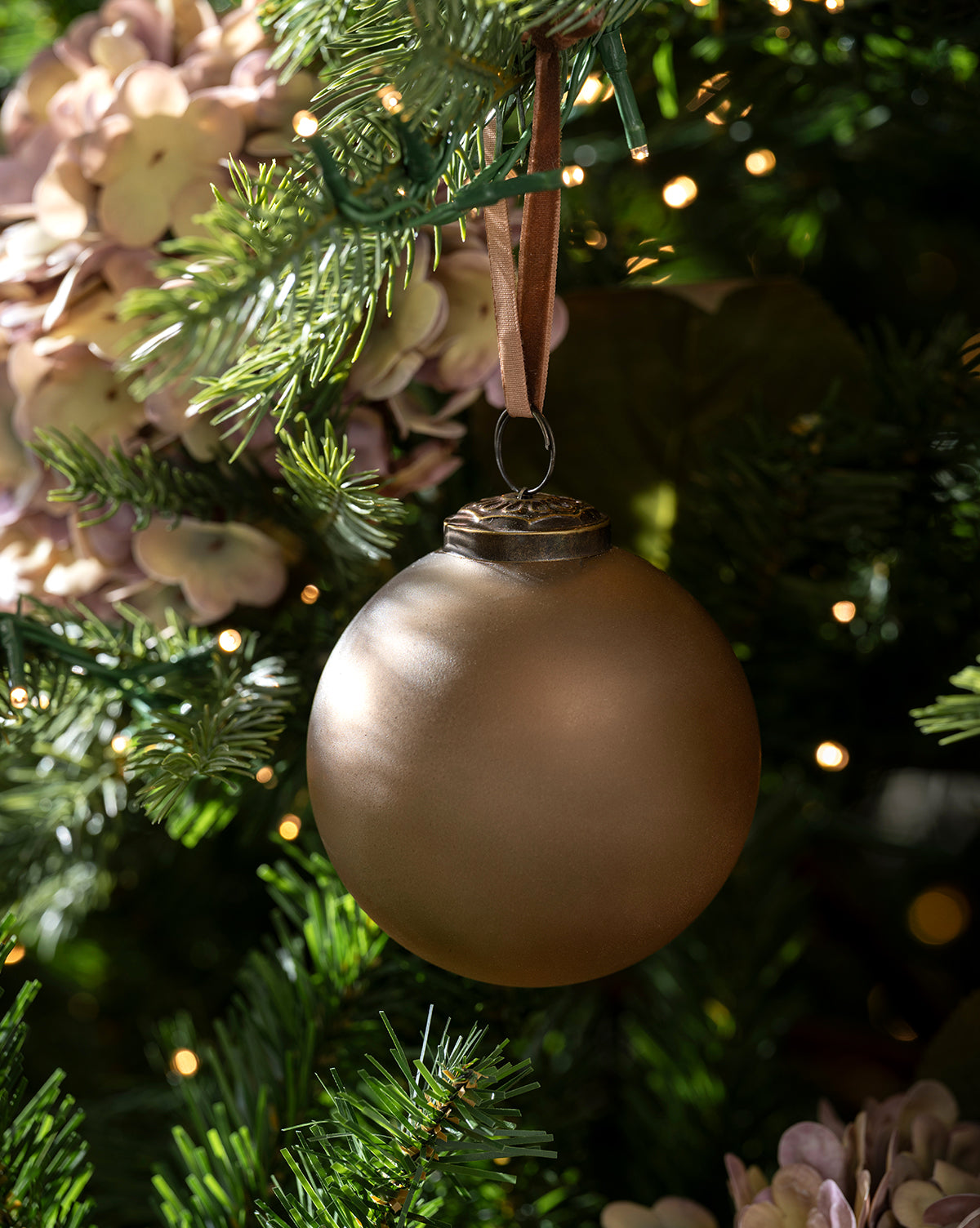 A matte gold ornament from the Noel Neutral Ornaments (Set of 9) hangs from a ribbon on a green Christmas tree, surrounded by pine needles, white flowers, and glowing lights—a perfect festive accent, by Vendixs