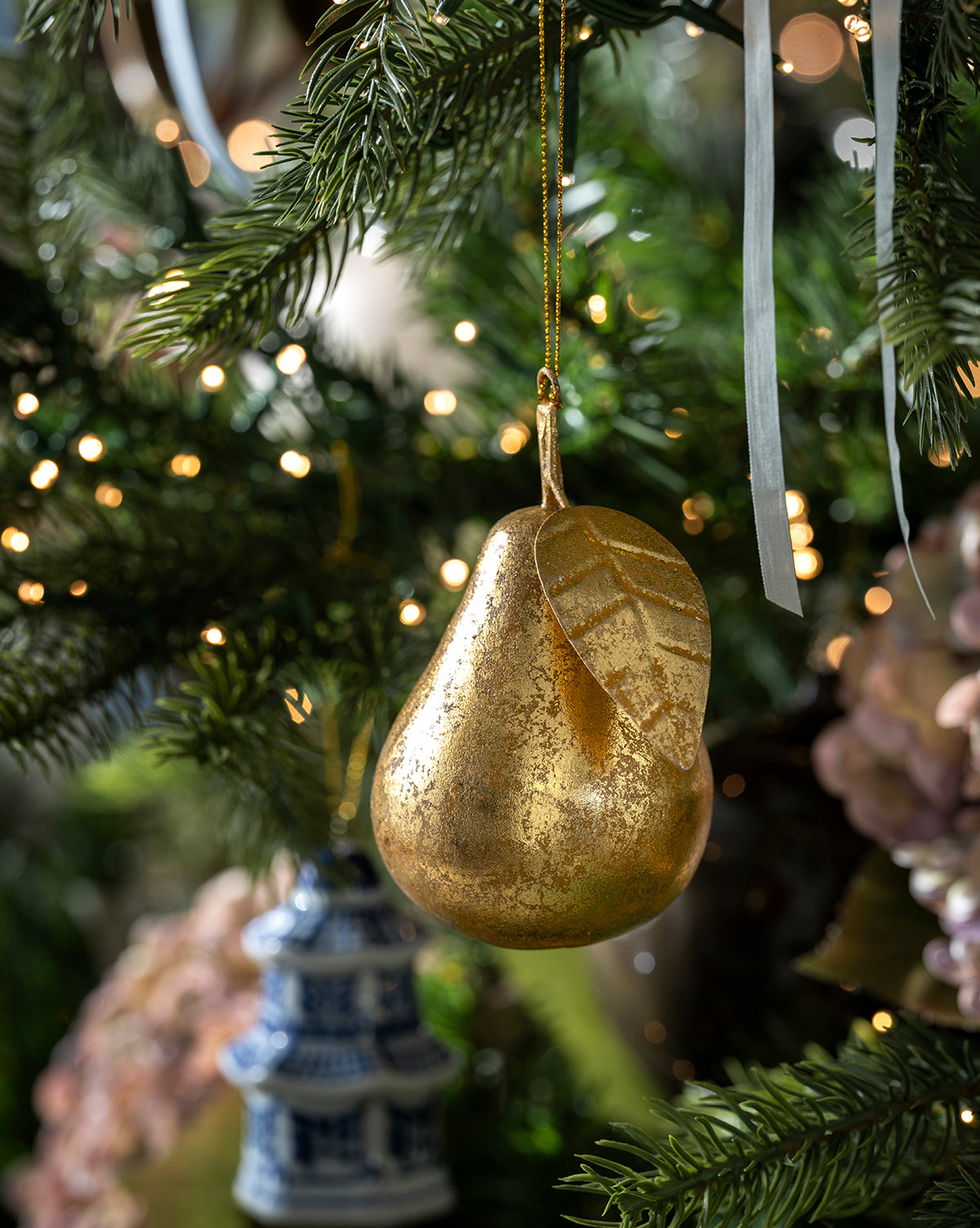 The Gilded Pear Ornament, featuring a leaf detail, hangs on a Christmas tree among pine needles and warm white lights, capturing the nostalgic charm of a vintage holiday decoration, by Vendixs
