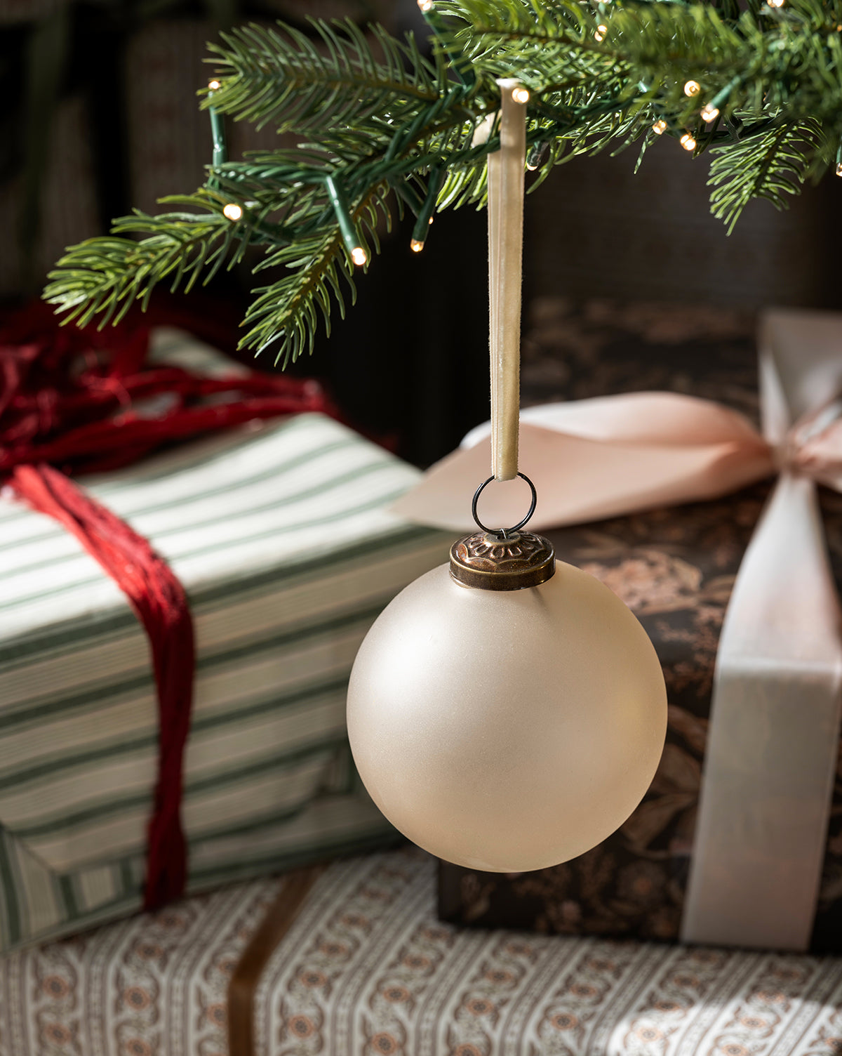 A Noel Neutral Ornament from the set of 9 hangs from a ribbon on a tree branch, with wrapped gifts in striped and patterned paper and festive ribbon visible in the background, by Vendixs