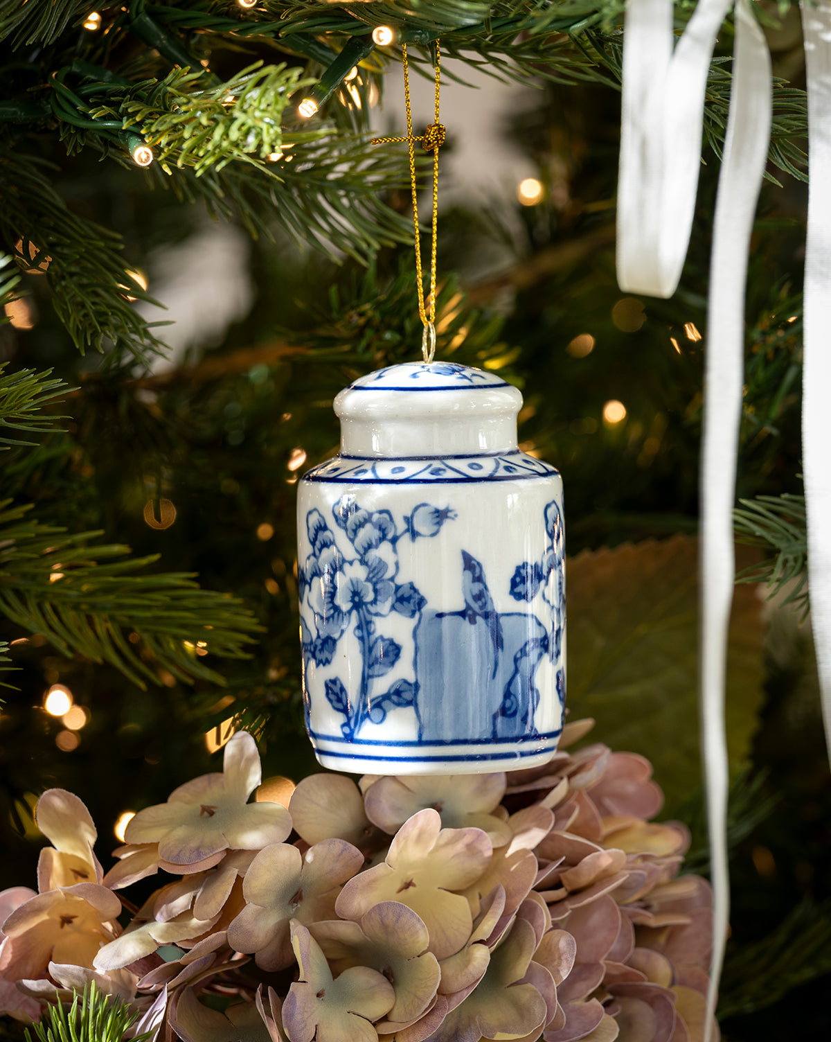 A blue and white floral porcelain jar ornament hangs on the Chateau Noel Tree, surrounded by greenery, warm lights, pale pink hydrangeas, and a luxurious velvet tree skirt, by Vendixs