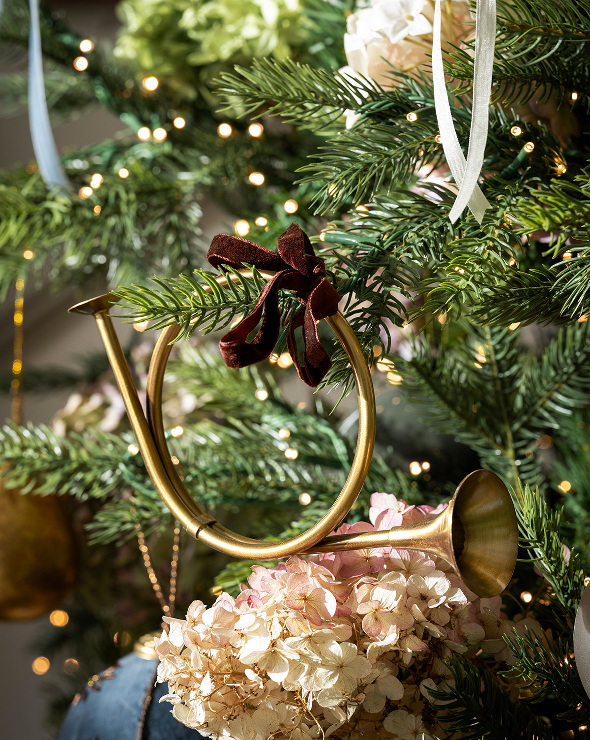 The French Horn Ornament, featuring a brass finish and red velvet bow, hangs on a holiday tree with green branches, white flowers, and twinkling string lights, by Vendixs