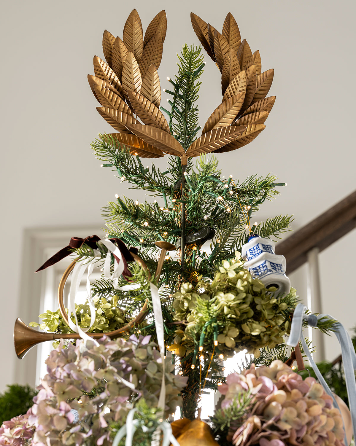 The top of this holiday tree is adorned with the Laurel Tree Topper, featuring a golden finish, green foliage, dried flowers, a blue-and-white teacup, ribbon, and a small golden trumpet, by McGee & Co.