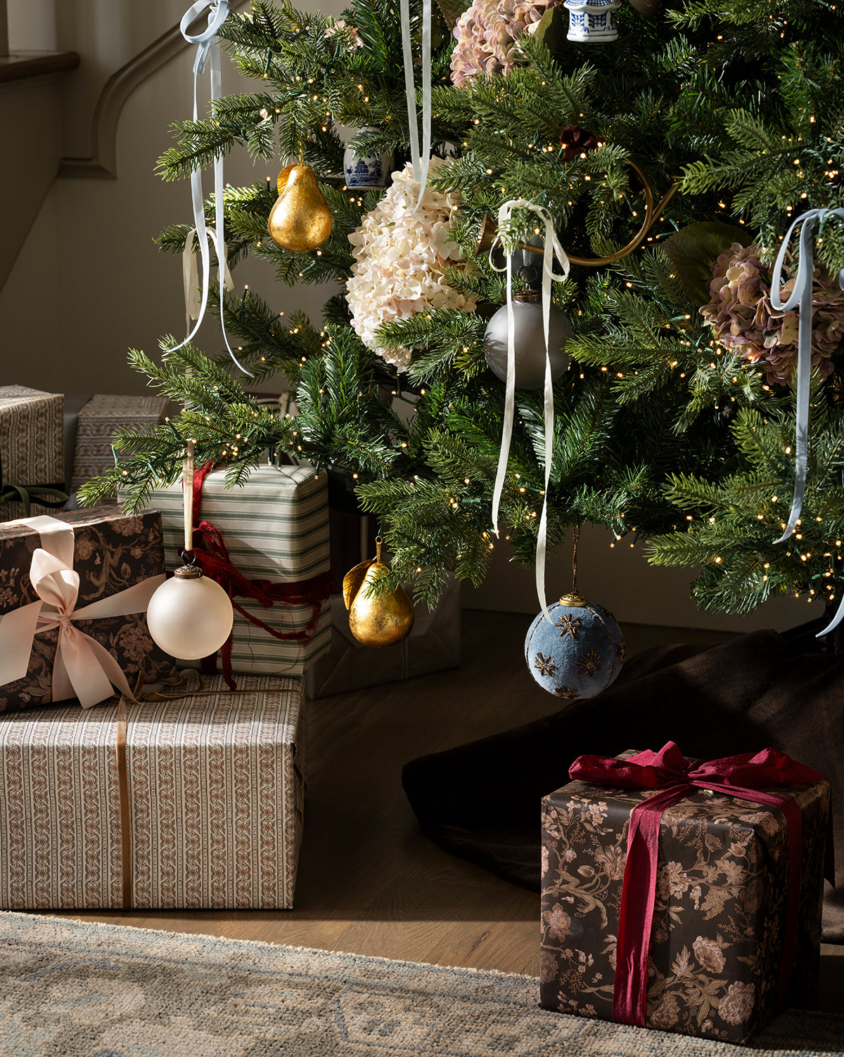 The Chateau Noel Tree, decorated with white and gold ornaments, ribbons, and fairy lights, stands above a velvet tree skirt with several wrapped presents and bows arranged nearby, by Vendixs