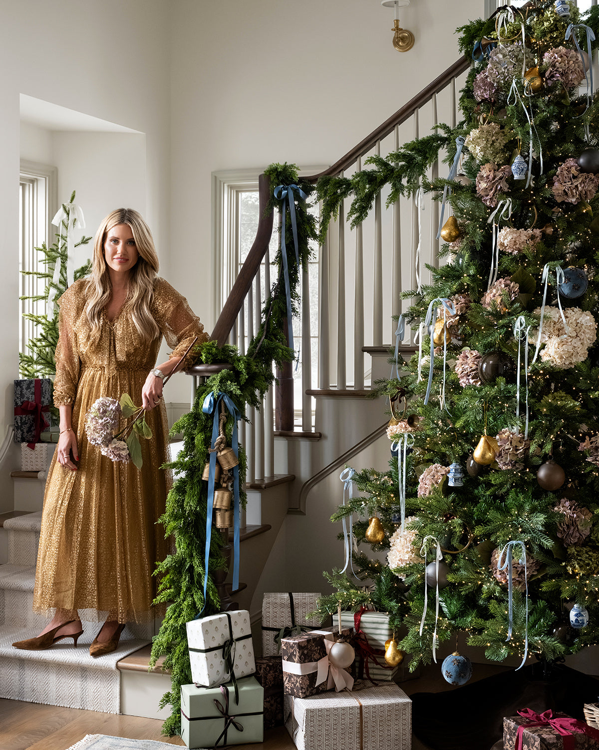 A woman in a gold dress stands by a staircase draped with garland, beside the Chateau Noel Tree decorated with ornaments and ribbons. Wrapped gifts sit on a velvet tree skirt in the bright, festive room, by Vendixs