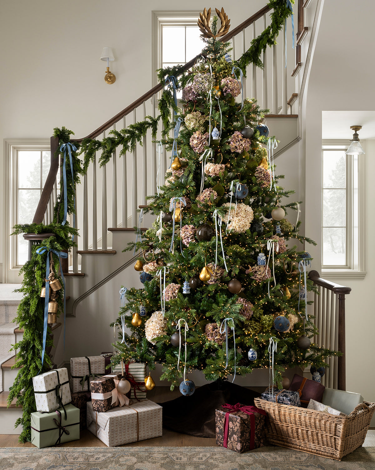 A 9' Norway Spruce Pre-Lit Faux Tree stands by the staircase decorated with garland. Wrapped presents and a basket rest beneath the tree, which is trimmed with ornaments, ribbons, and dried flowers, as sunlight pours in through nearby windows, by Vendixs