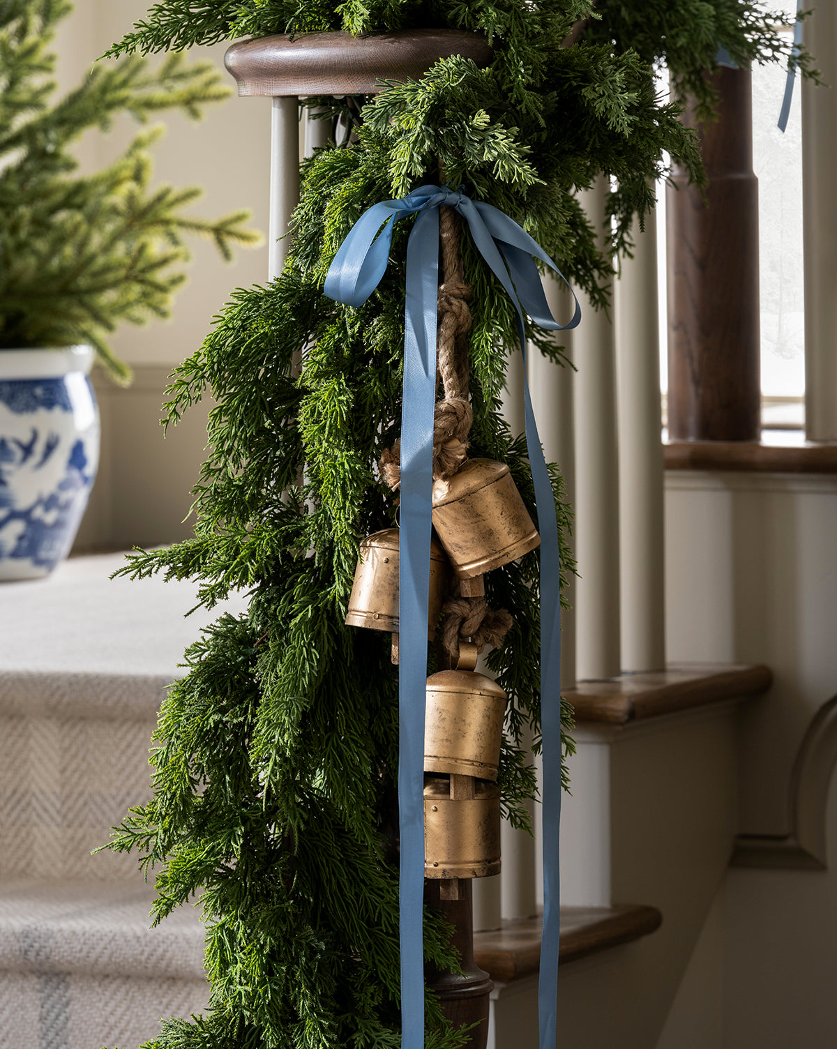 A staircase is adorned with lush green garland, gold Hanging Iron Bells, and a blue ribbon. In the background, a blue and white pot holds a small evergreen tree, creating festive holiday décor, by Vendixs