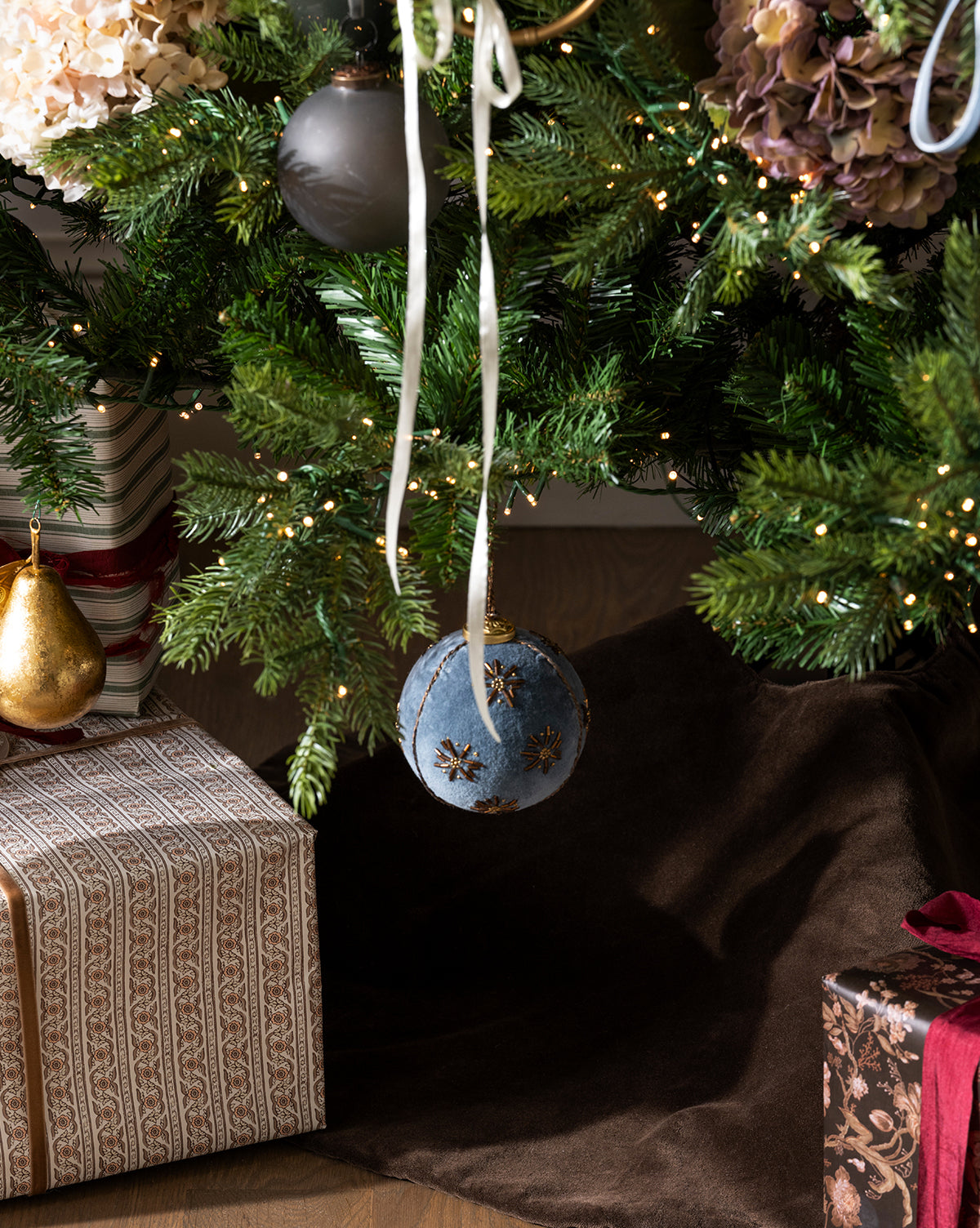A decorated Christmas tree with a blue ornament and white ribbon hangs above wrapped gifts, all set atop the Velvet Brown Tree Skirt, which adds a touch of luxury and elegance to your holiday decor, by Vendixs