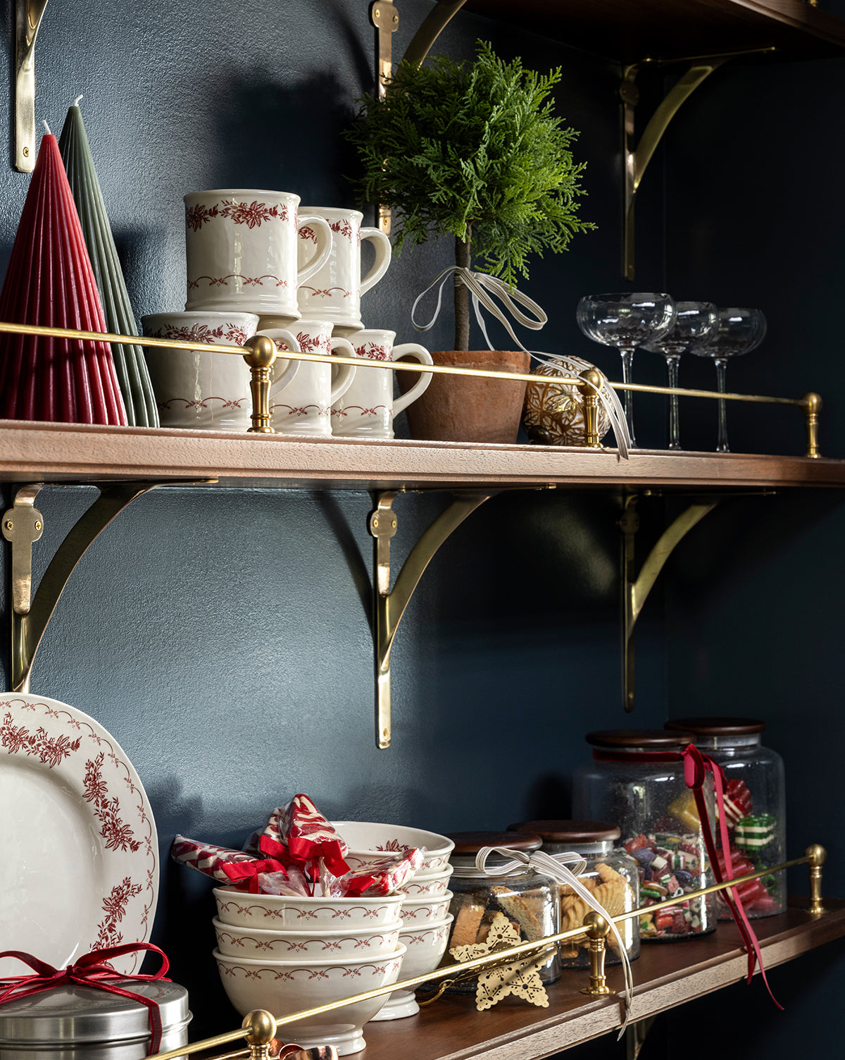 Wooden shelves with gold brackets hold stacked red-patterned mugs, bowls, plates, wine glasses, wrapped cookies, the 15.5" Cypress Faux Potted Topiary in a terracotta pot, red candles, and festive kitchen decor against a dark blue wall, by Vendixs