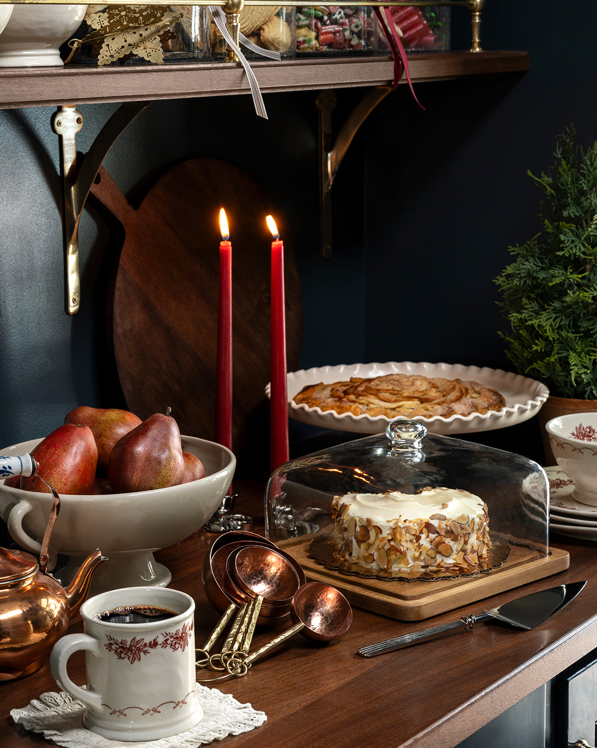 A cozy holiday tablescape showcases a cream almond cake under glass, a bowl of pears, copper kitchenware, a teapot, cups, pastry on a plate, and two red candles in the elegant Silver Bow Taper Holder, by Vendixs