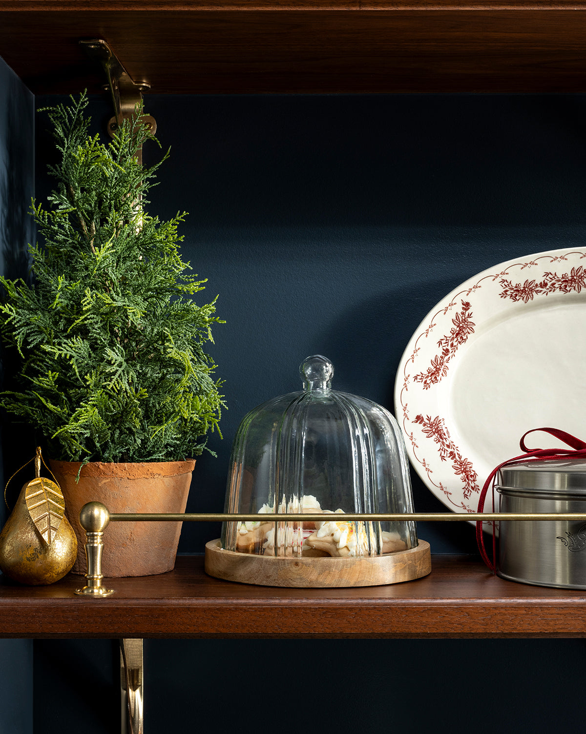 A 17" Cypress Faux Potted Tree, a golden pear ornament, a cheese plate with glass dome, a red-ribboned tin, and a white platter with red border are arranged on a wooden shelf against a dark wall, by Vendixs