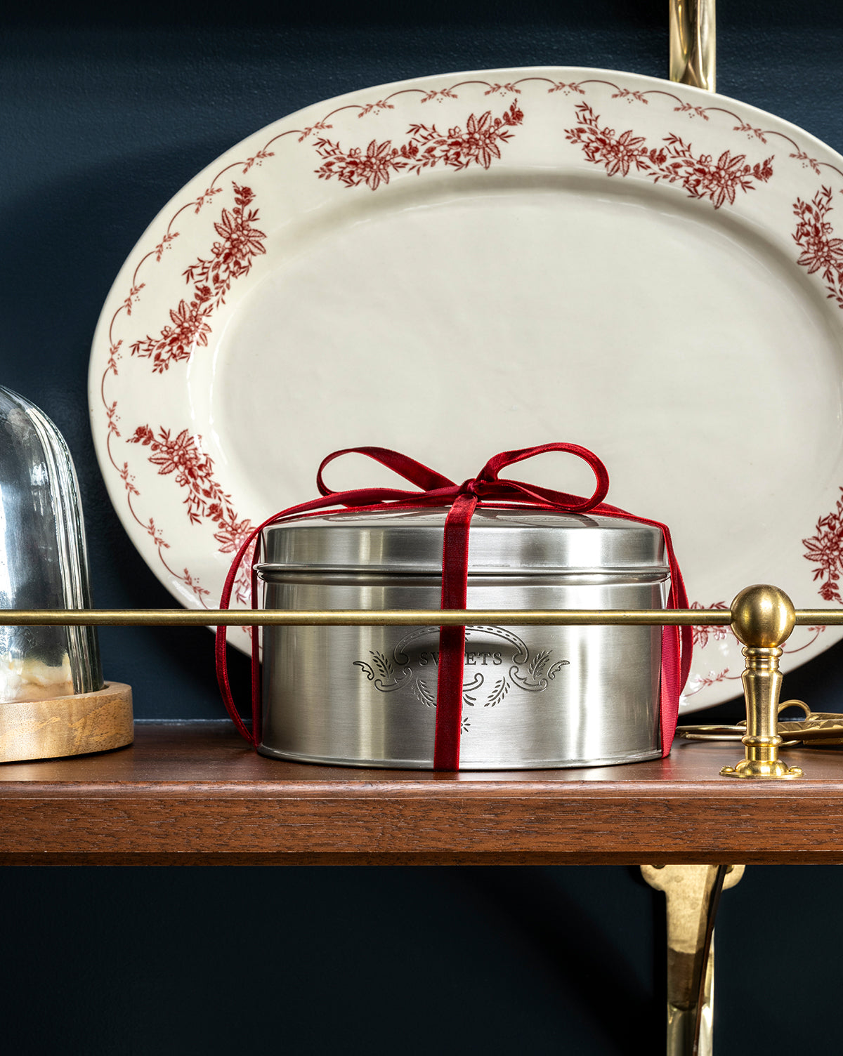 The Sweets Cookie Tin, silver with engraved details and a red velvet ribbon, brings vintage charm to a wooden shelf beside a white platter with red floral trim. A glass dome and brass supports complete the elegant display, by Vendixs