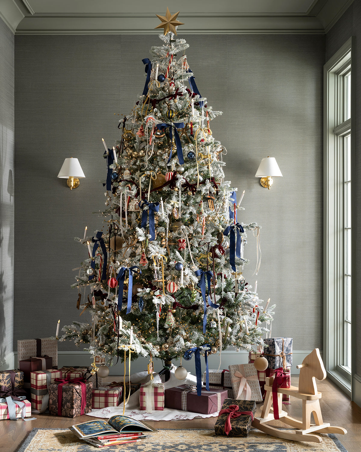 A 9' Snowy Grandis Fir Pre-Lit Faux Tree, decorated with ribbon, ornaments, and a gold star, stands in the living room with wrapped gifts beneath. Sunlight streams through large windows as two wall sconces frame the festive tree, by Vendixs