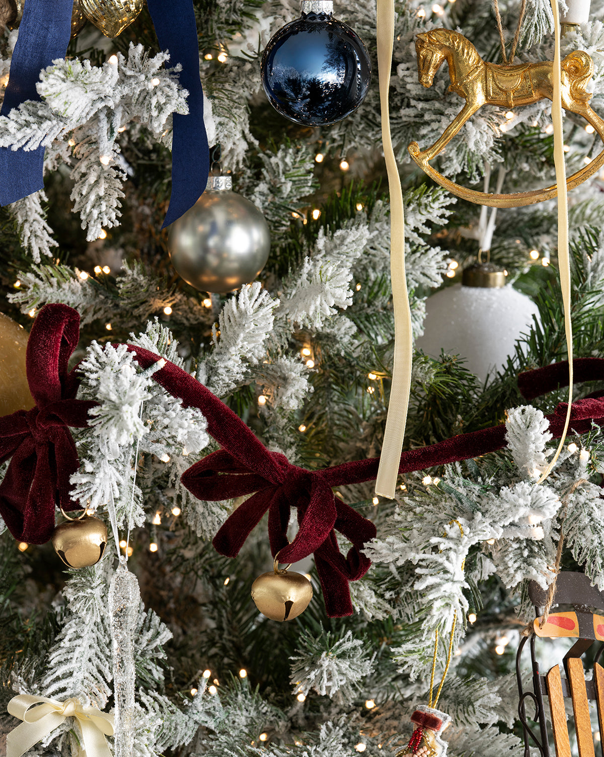 Close-up of a Christmas tree adorned with snow-dusted branches, gold and silver ornaments, a gold rocking horse, blue and burgundy velvet bows, the 6' Mini Bows & Bells Garland, jingle bells, and white string lights for festive decor, by McGee & Co.