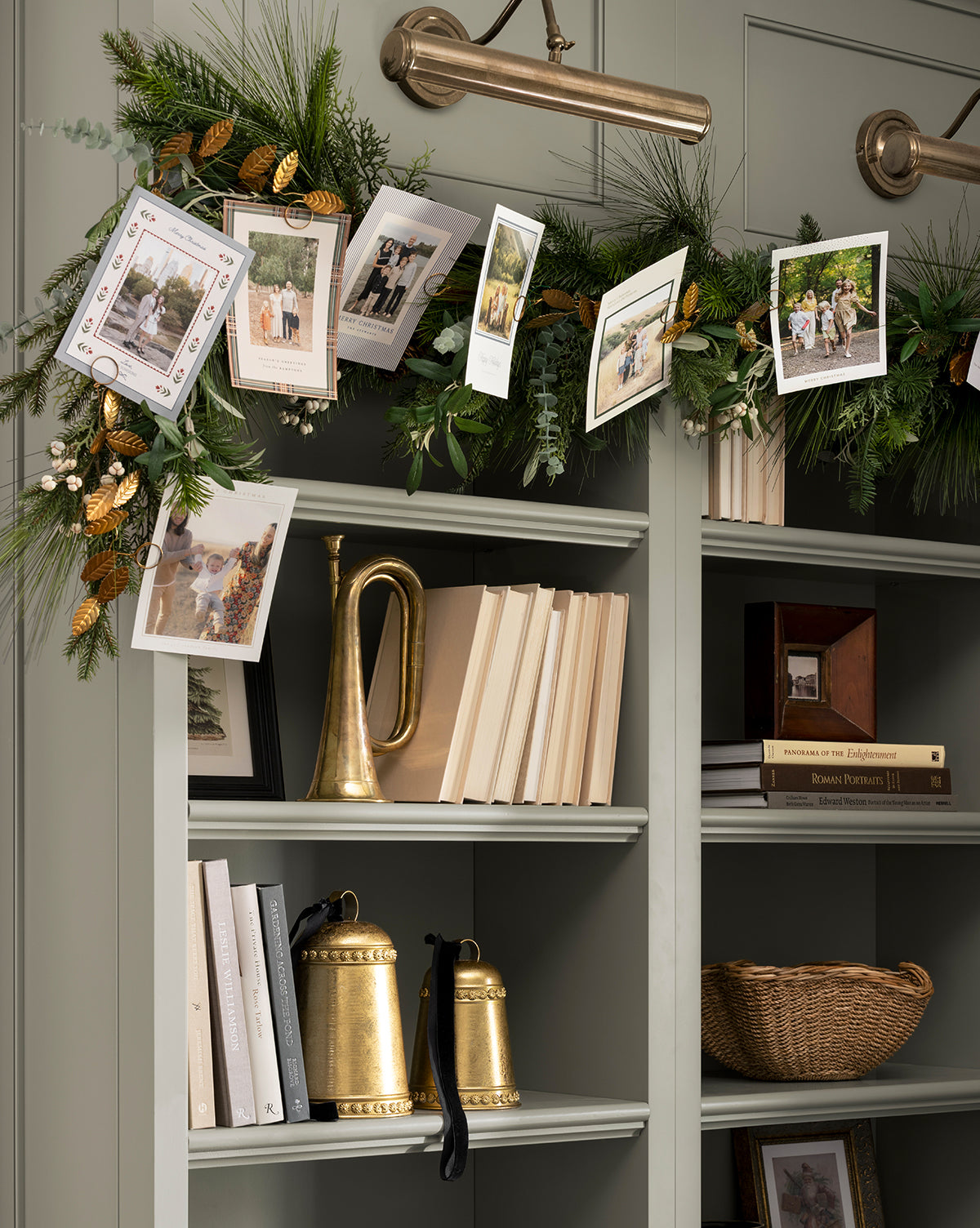 A 6' Card Holder Garland with pine and gold accents hangs above bookshelves, displaying holiday cards. The shelves feature books, a gold horn, gold bells, and a woven basket for a festive and cozy atmosphere, by Vendixs