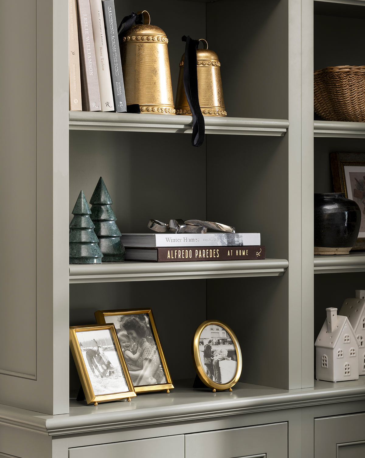 A bookshelf styled for the holidays features gold cowbells, stacked books, framed black-and-white photos, a round clock, a black vase, a wicker basket, a white ceramic house, and the Jolee Mantel Trees (Set of 3) as festive green accents, by Vendixs
