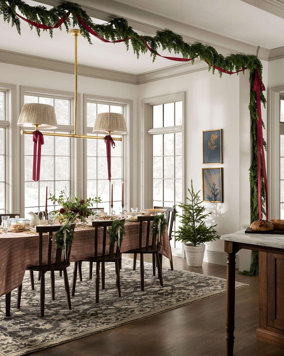 A dining room features a 6' Cedar Pre-Lit Faux Garland, red ribbons, a festive table, two small potted Christmas trees, and large windows streaming natural light—perfect for holiday home decor, by Vendixs