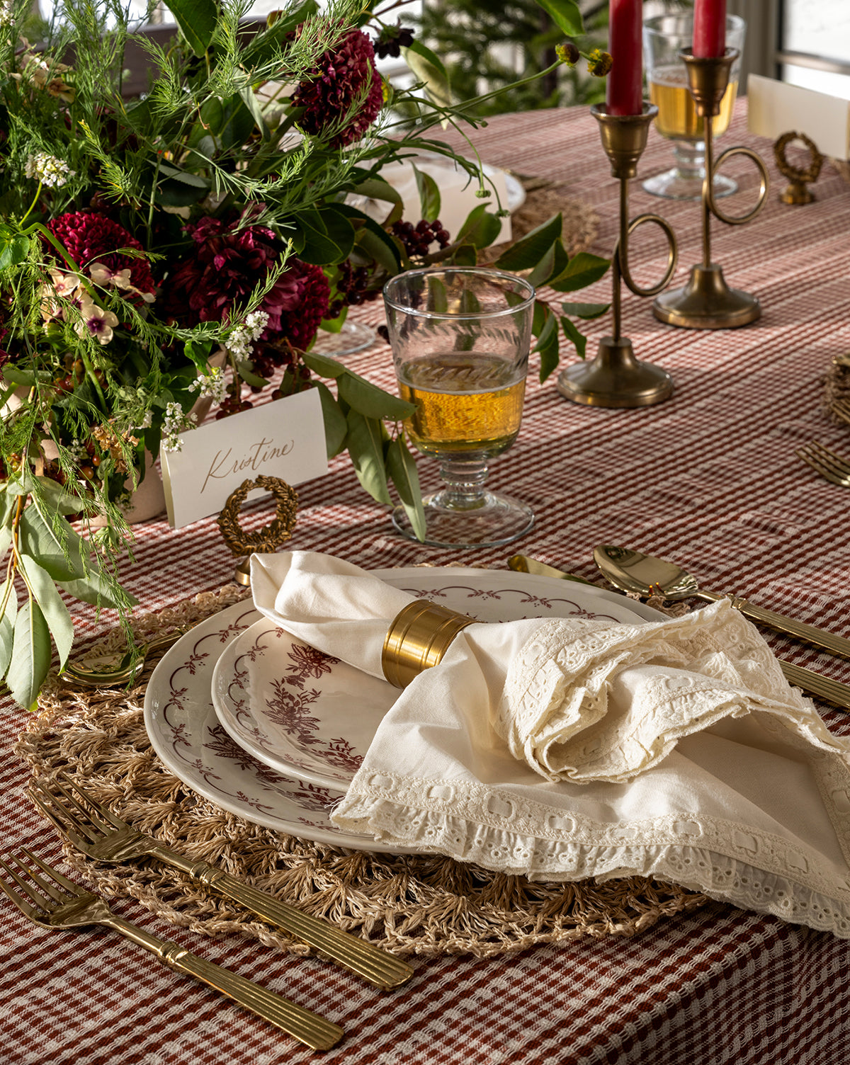 Elegant holiday table with vintage plates, lace napkin in an Argent Napkin Ring (Set of 4), woven placemat, gold cutlery, amber drinkware, red candles, floral centerpiece, name card “Kristine,” and a checkered tablecloth, by Vendixs