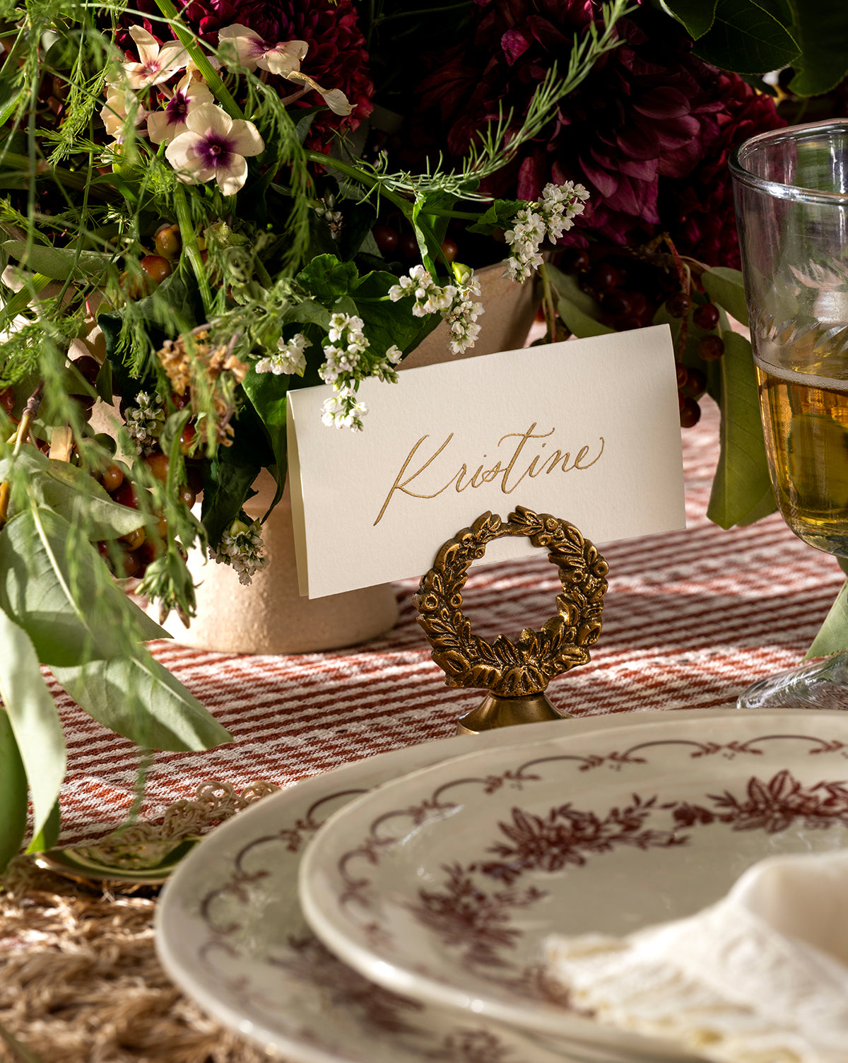 A table setting features floral-patterned plates, a glass of white wine, and a handwritten place card reading Kristine in the Laurel Place Card Holder (Set of 4), with lush holiday floral arrangements in the background, by Vendixs