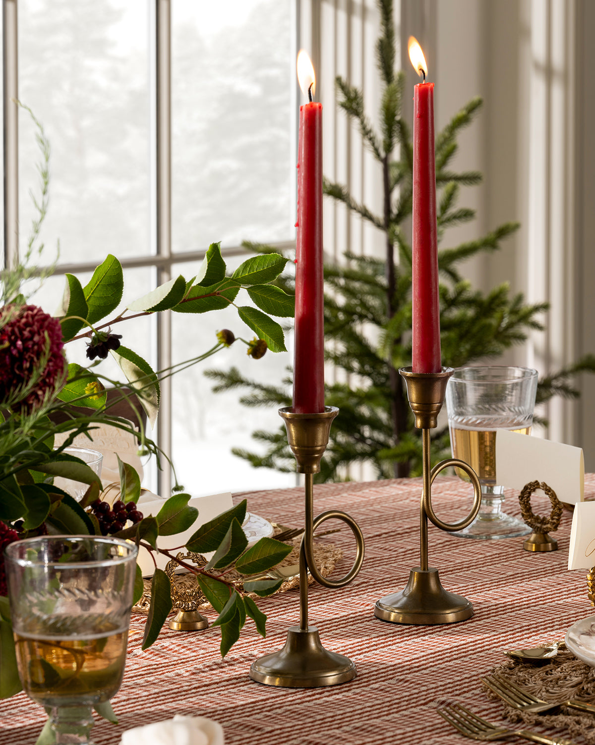 Two lit red taper candles in French Horn Taper Holders sit on a patterned tablecloth beside green foliage, a wine glass, and holiday decor, with festive styling and a window with a small Christmas tree in the background, by Vendixs