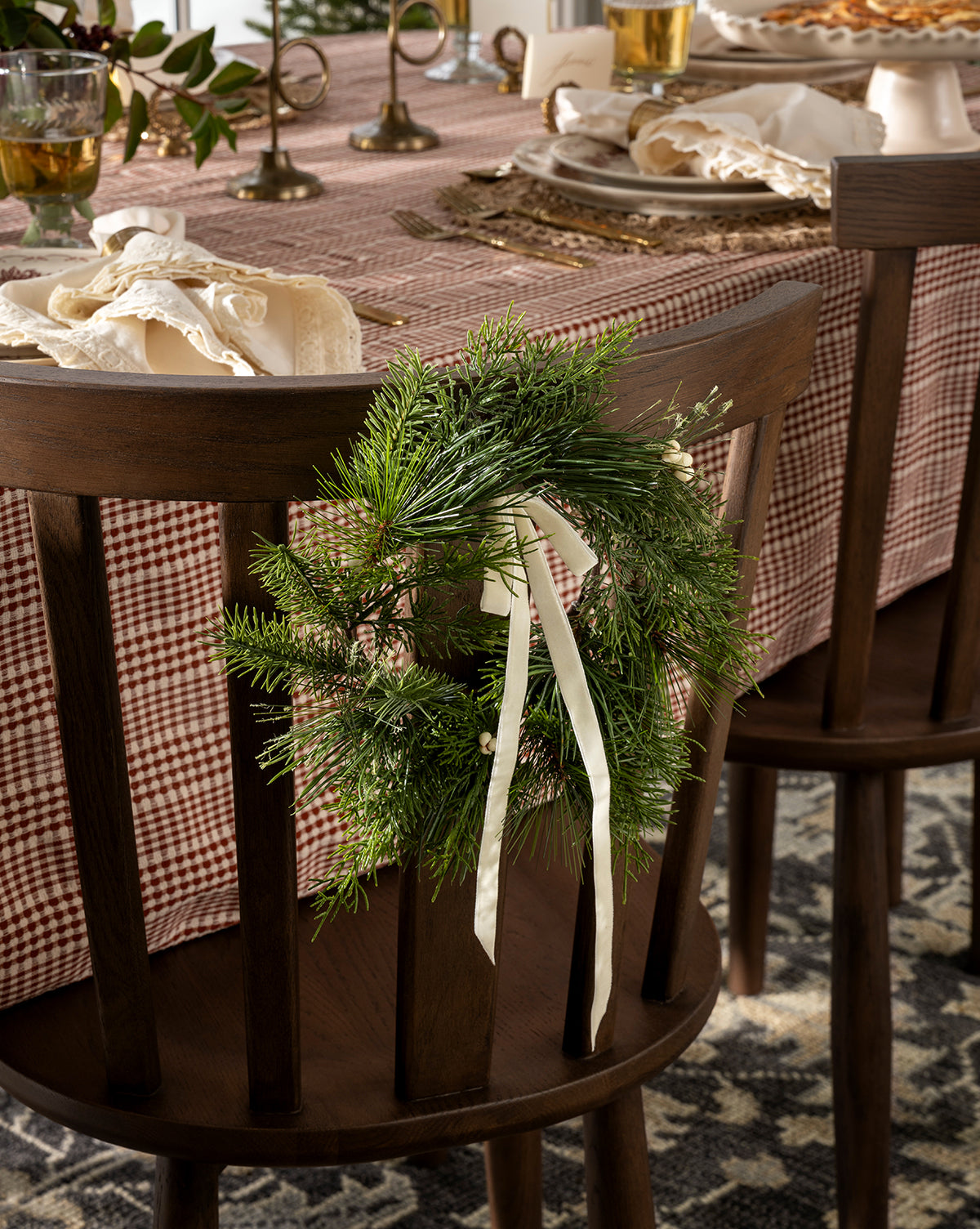 An 8" Pine Needle & Berry Faux Wreath with a white ribbon hangs on a dark wooden chair, bringing festive cheer to a dining table set with plaid linens, napkins, plates, glasses, and candles, by McGee & Co.