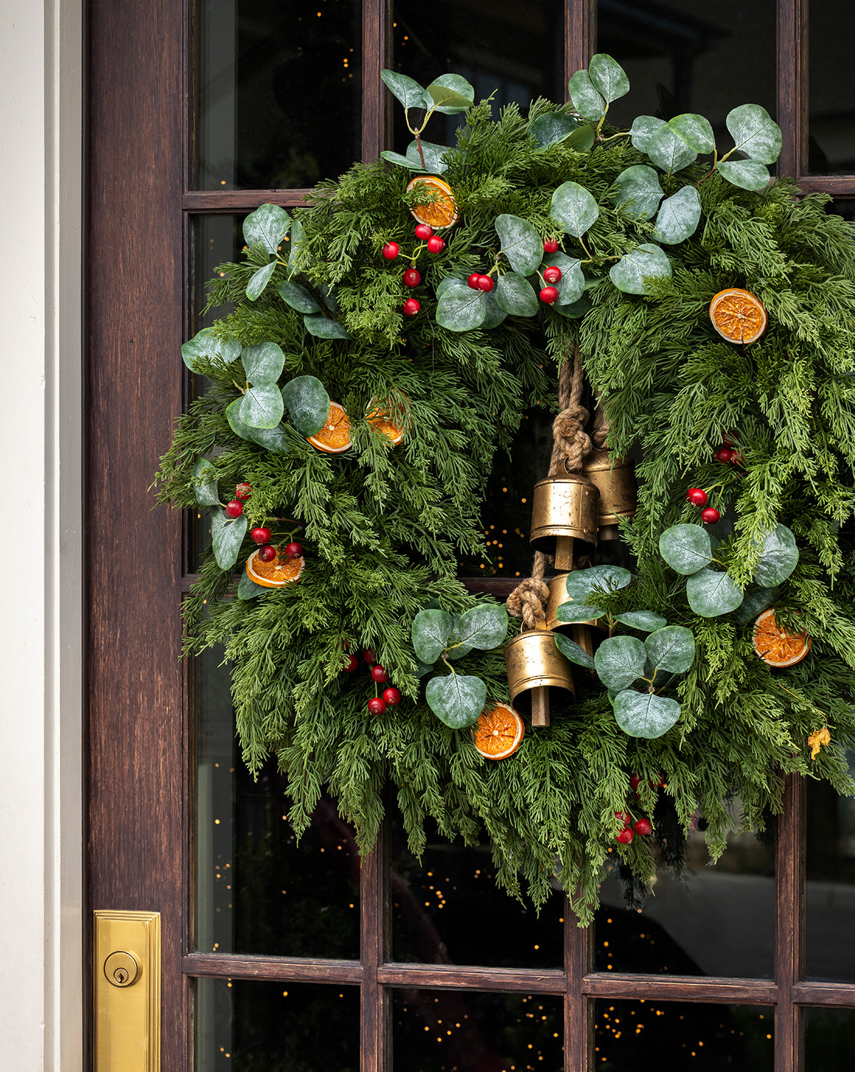 The 30" Orange & Eucalyptus Faux Wreath, featuring evergreen branches, eucalyptus leaves, red berries, dried orange slices, and gold bells, hangs on a dark wooden door with a brass handle, by Vendixs