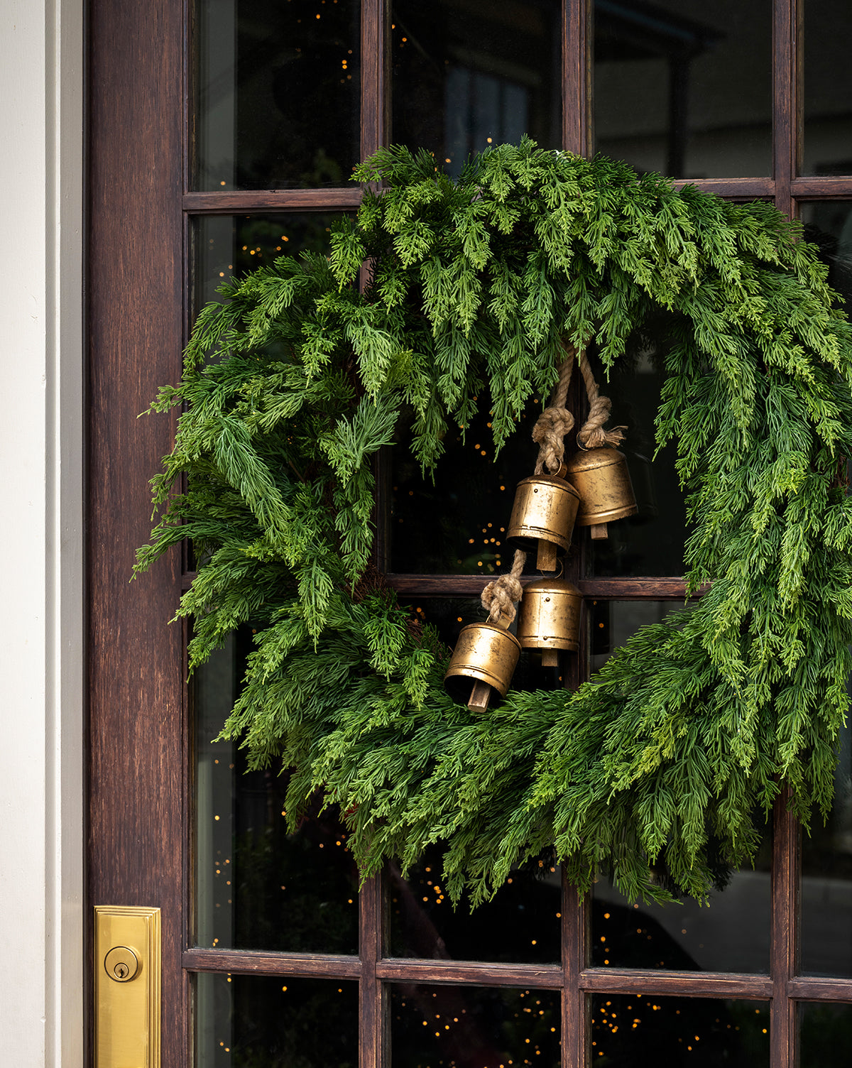 A 30" Classic Cedar Pre-Lit Faux Wreath with three brass bells hangs on a wood and glass door with a gold handle, as the glow from a Christmas tree inside creates festive Holiday Décor, by Vendixs