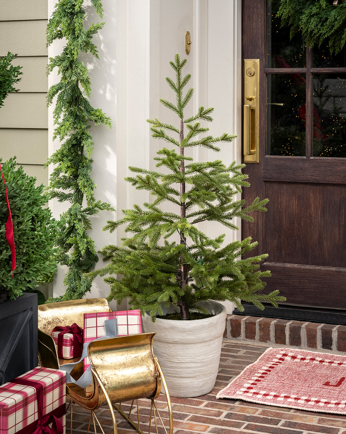 The Faux Spruce Pre-Lit Potted Tree stands on a brick porch by a dark wooden door, accented with wrapped gifts, a golden sleigh, festive garland, and a red plaid rug for a cozy holiday ambiance, by Vendixs