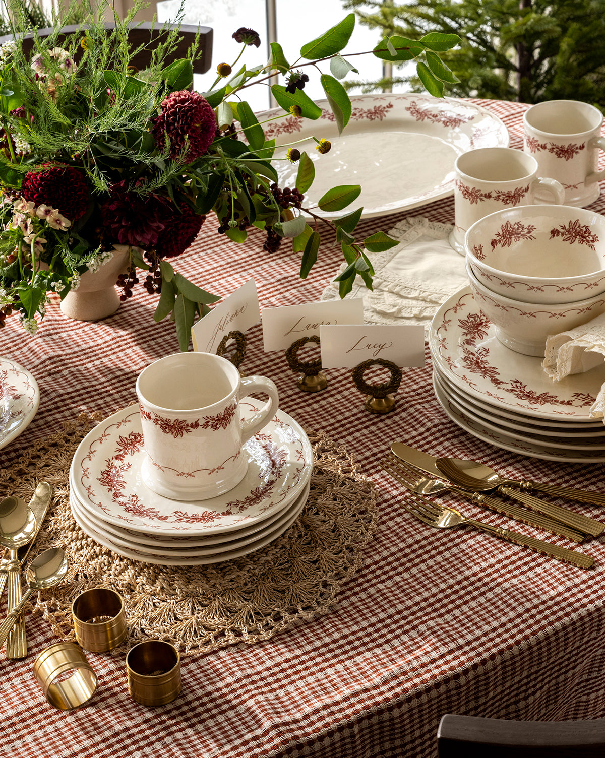 A holiday table set for a meal with vintage-style red floral china, gold cutlery, Argent Napkin Rings (Set of 4), woven placemats, a floral centerpiece, place cards, and a checkered tablecloth, by Vendixs