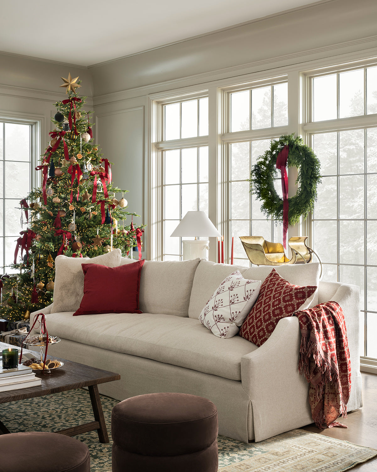 A cozy living room features a white sofa with red and white pillows, festive décor, a Christmas tree with ornaments and ribbons, and a 30" Classic Cedar Pre-Lit Faux Wreath on the window, illuminated by natural light, by Vendixs