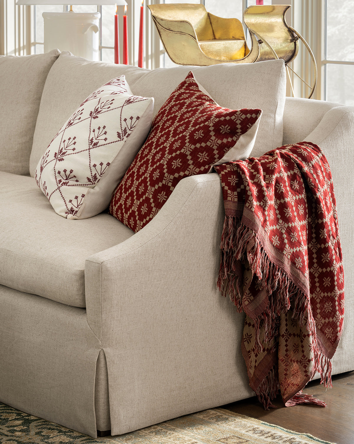 A beige sofa with two patterned throw pillows—one white with red embroidery, one red with a white geometric design—and the Jude Reversible Woven Throw draped over the armrest, by Vendixs