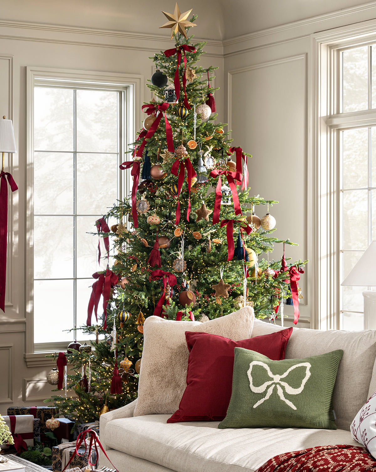 A Christmas tree with red ribbons, ornaments, and a gold star stands by large windows. Wrapped gifts sit below, while a beige sofa topped with red and green pillows plus the Bella Pillow Cover adds cozy holiday charm, by Vendixs