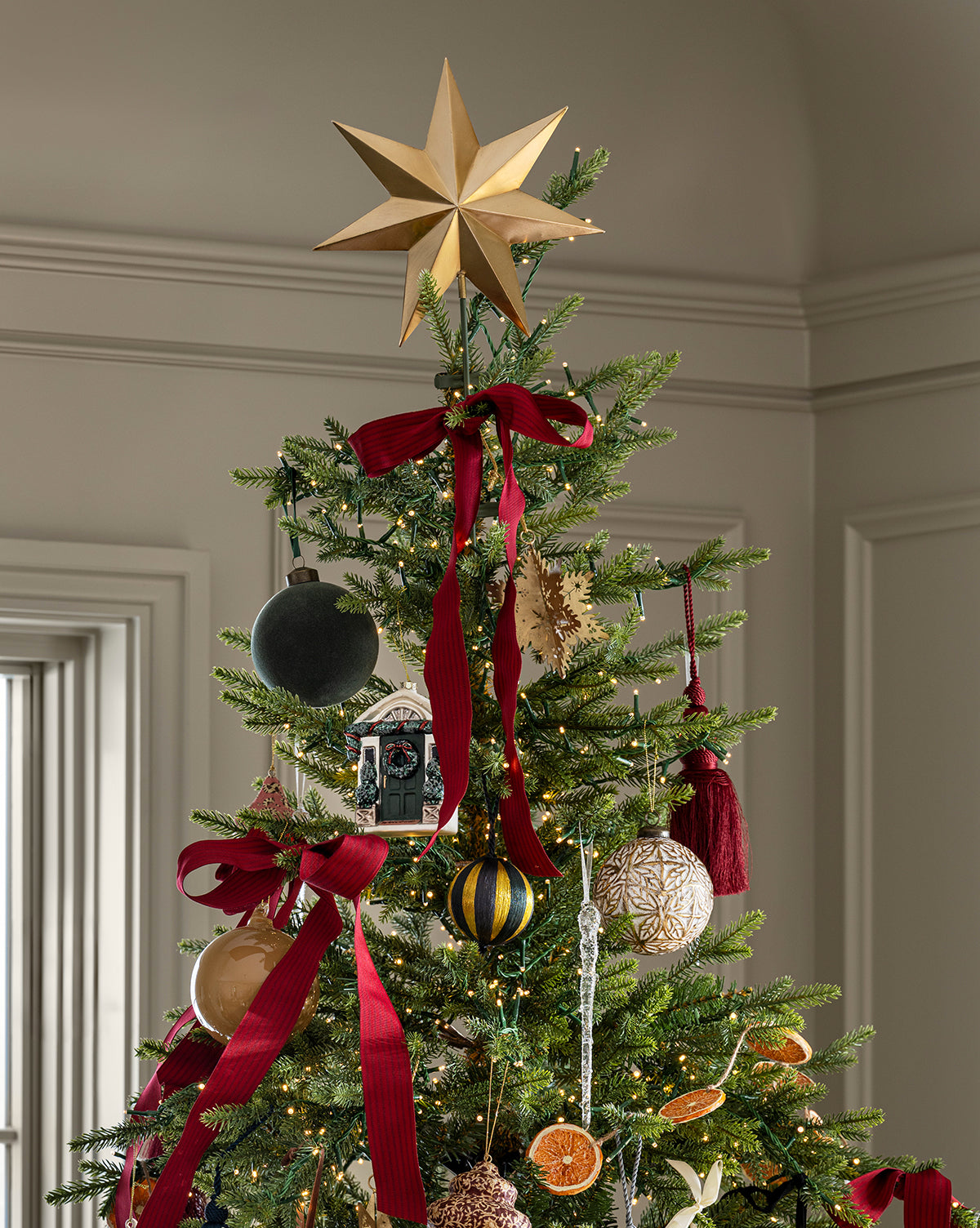 The Classic Christmas Tree is topped with a gold star and decorated with red ribbons, ornaments, dried orange slices, and festive touches. Its setting features light-colored walls and trim for a cozy holiday feel, by Vendixs