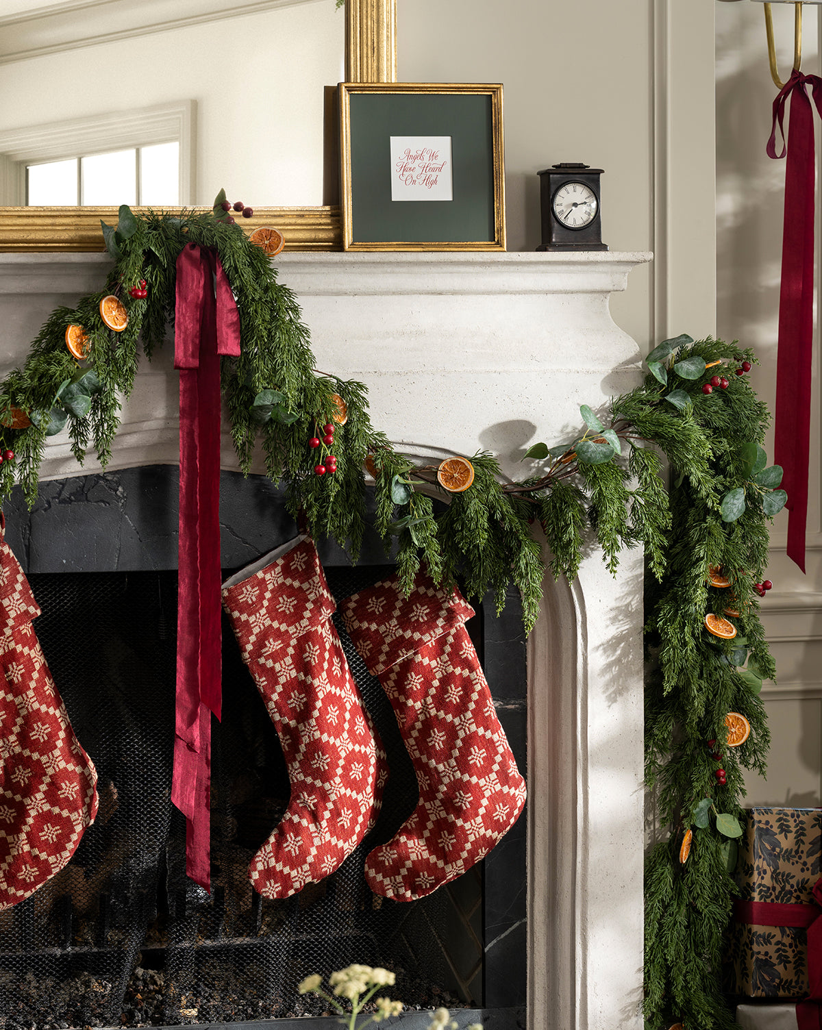 A festive holiday mantel features green garland, dried orange slices, and red bows. Three Jude Stockings with woven texture hang from the mantel, while a framed picture and small clock sit above, by Vendixs