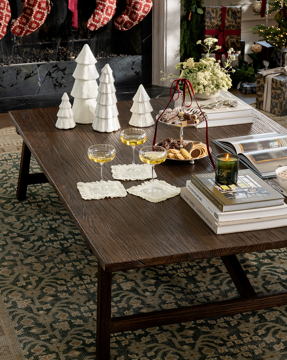 A wooden coffee table decorated for the holidays with handcrafted white ceramic trees, a Confectioners Paper Mache Tree, three champagne glasses on coasters, cookies on a tiered tray, books, a green candle, and festive décor by the fireplace, by Vendixs
